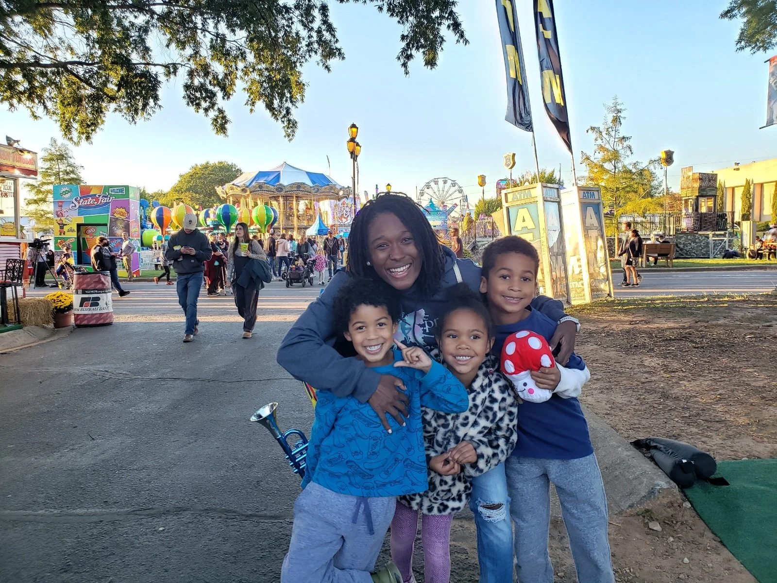 A group of four children and a woman, all smiling and embracing each other, at a carnival or fair with rides, balloons, and people in the background.