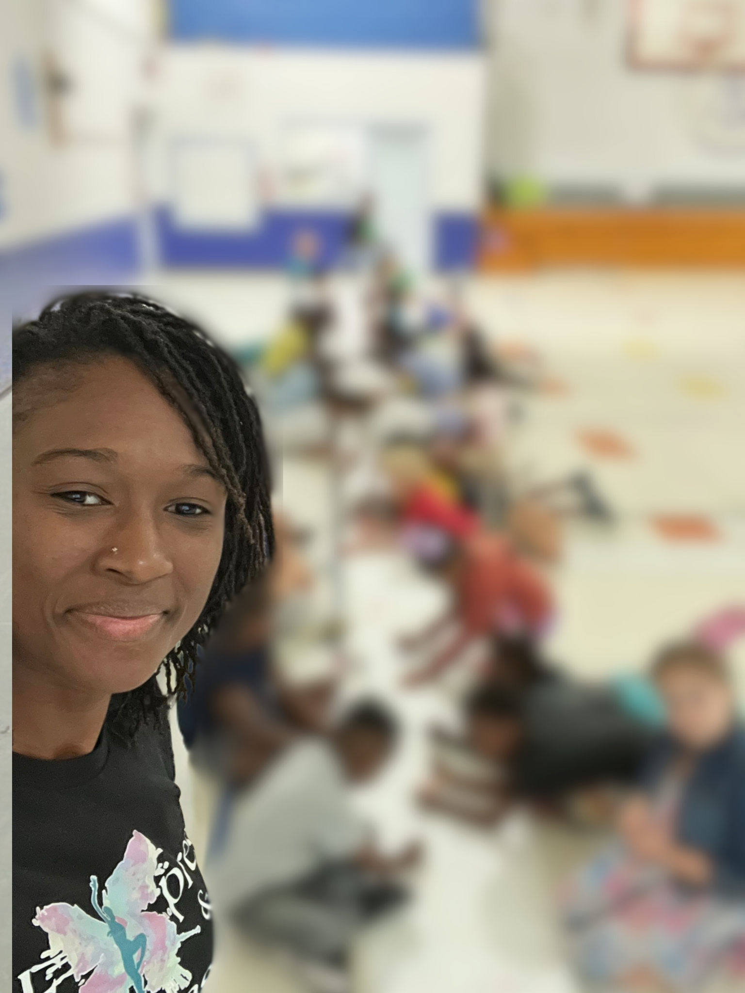 A woman with braided hair smiling at the camera in a classroom with children sitting on the floor in the background.