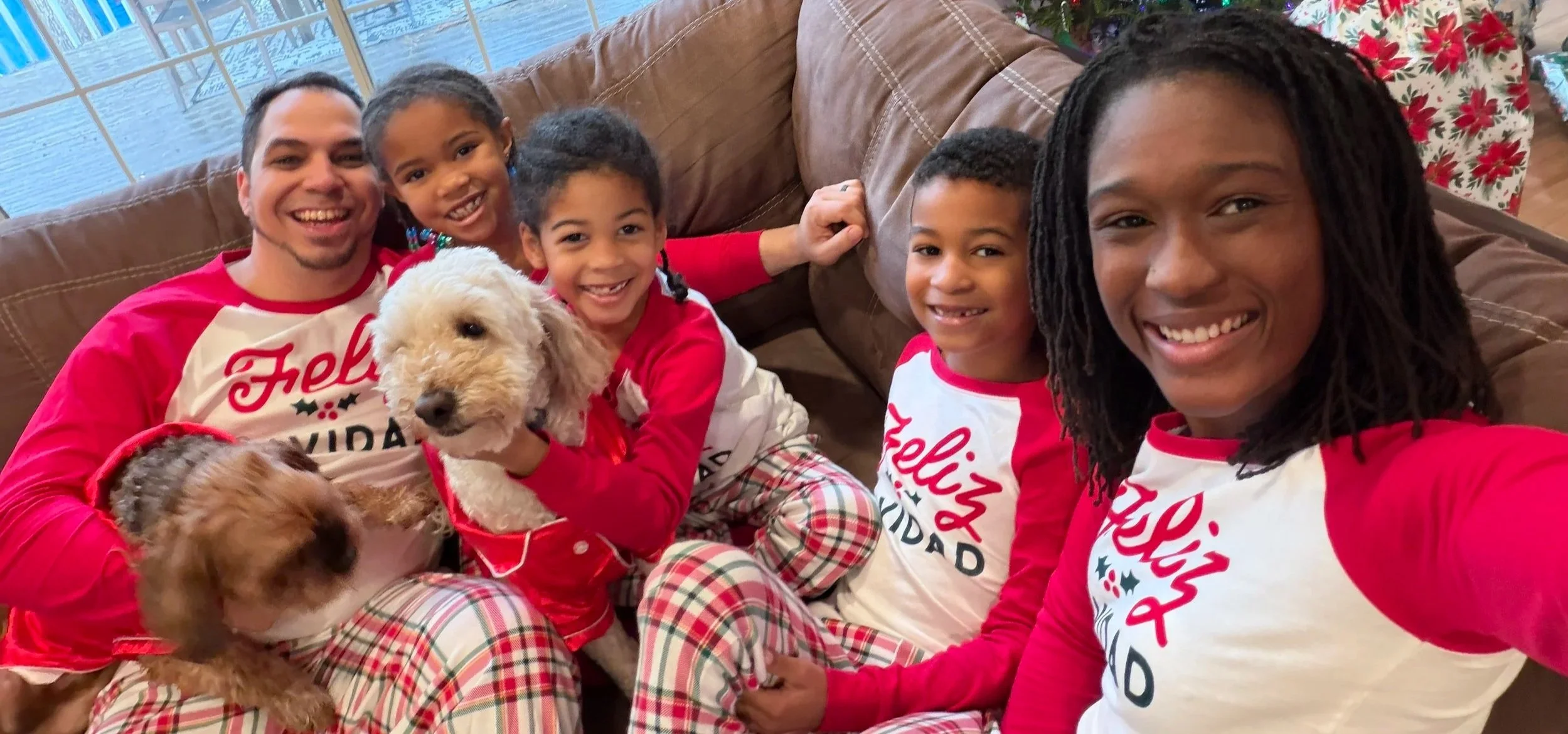 Family celebrating Christmas in matching holiday pajamas, including a man, three girls, and two dogs on a brown couch, with Christmas pajamas and a window in the background.