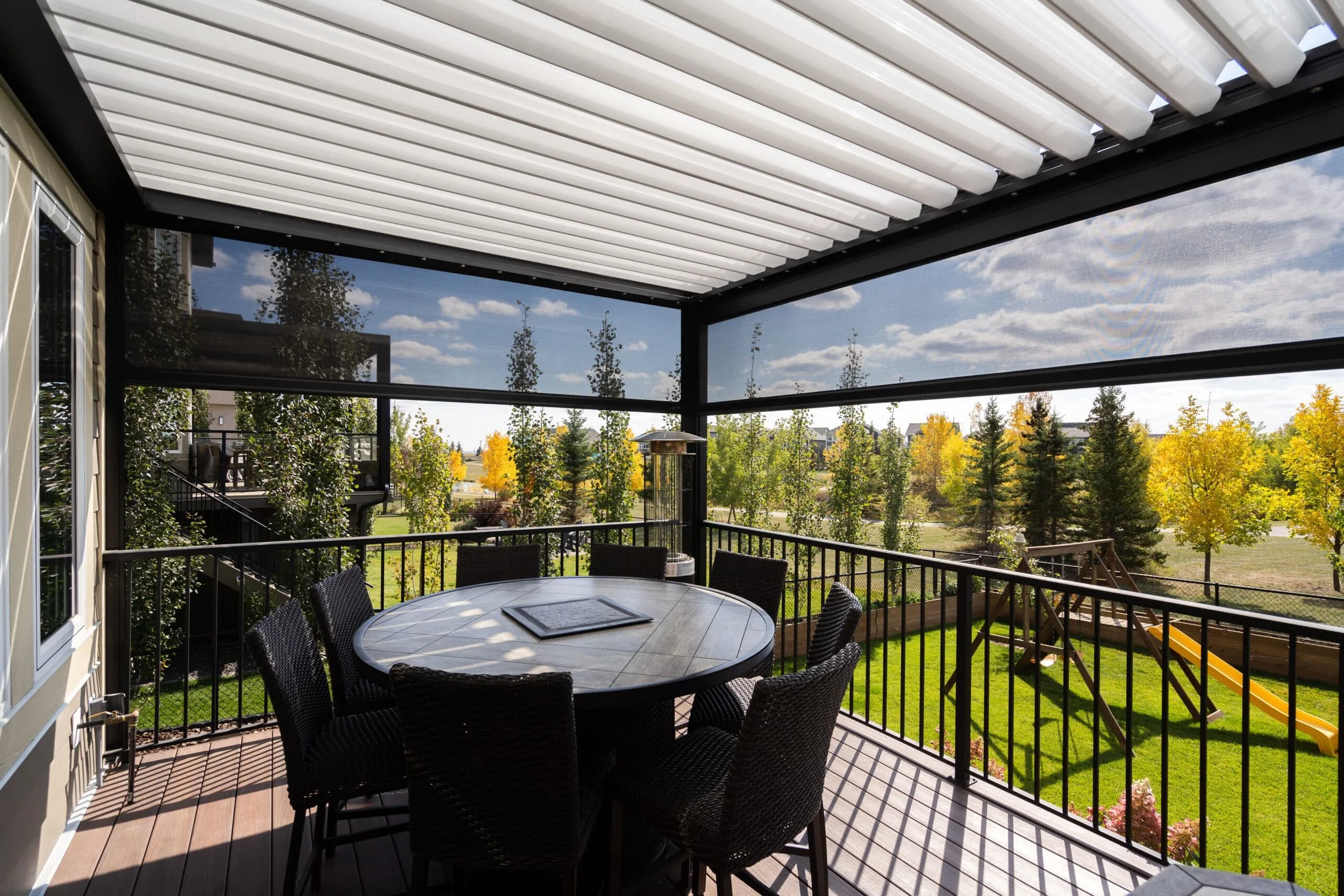 Covered patio with a round dining table and six chairs overlooking a backyard with trees, a children's slide, and a grassy area under a partly cloudy sky.