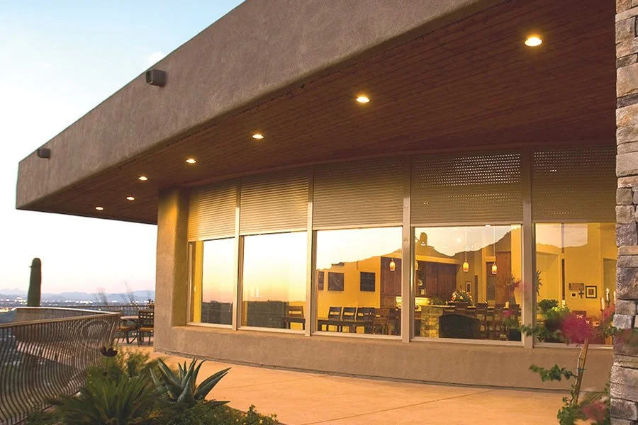 Modern building with large glass windows reflecting interior lights and sunset, exterior patio with plants and outdoor seating, cityscape in the distance.