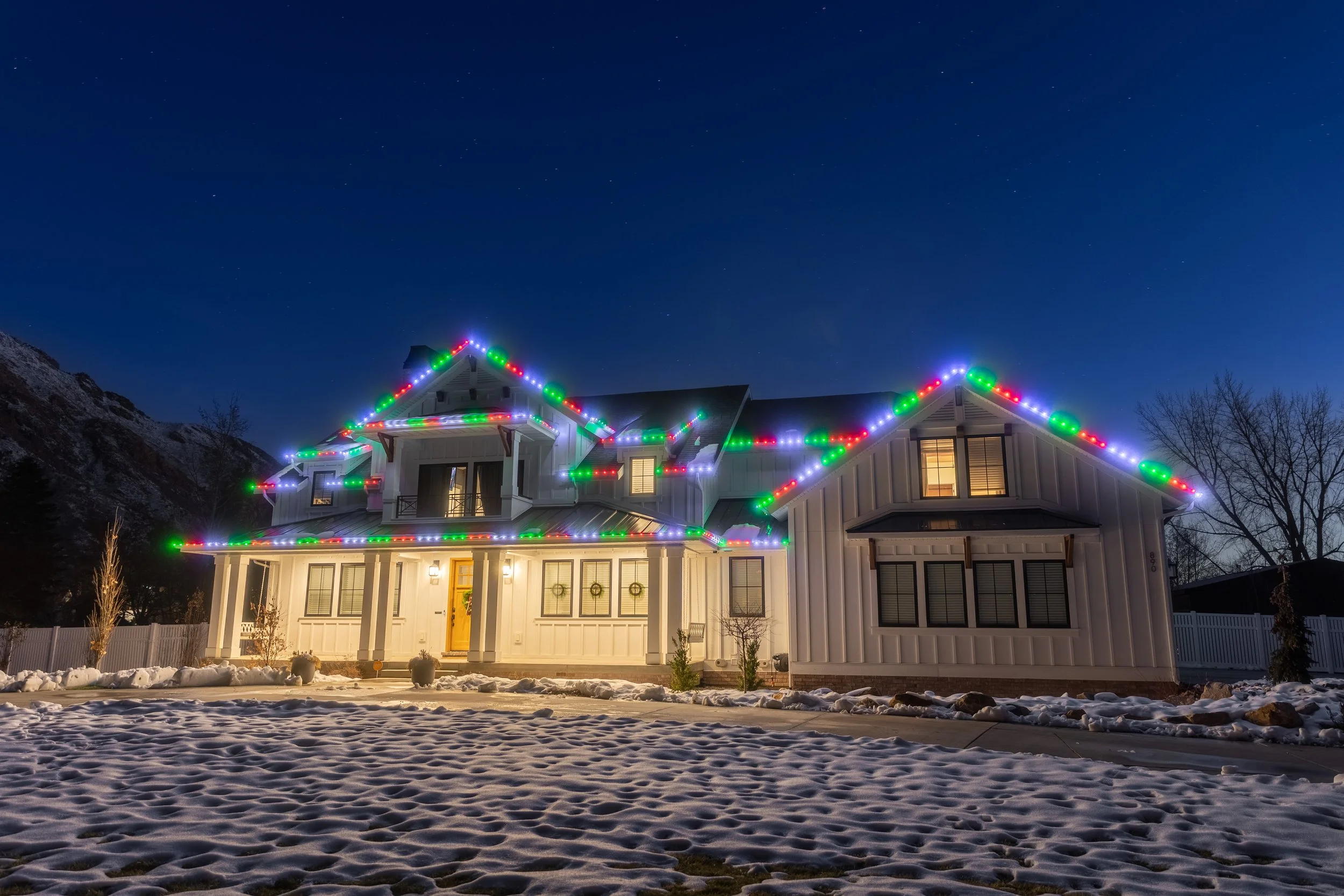 A house decorated with colorful Christmas lights at night, with snow on the ground and a mountain in the background.