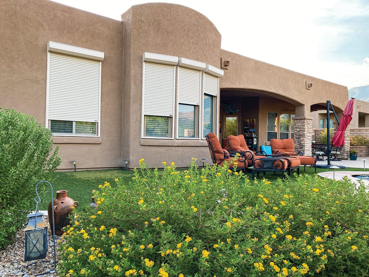 Backyard patio with outdoor seating, a large shrub with yellow flowers, a clay vase, a lantern, a barbecue grill, a red umbrella, and a mountain in the background.