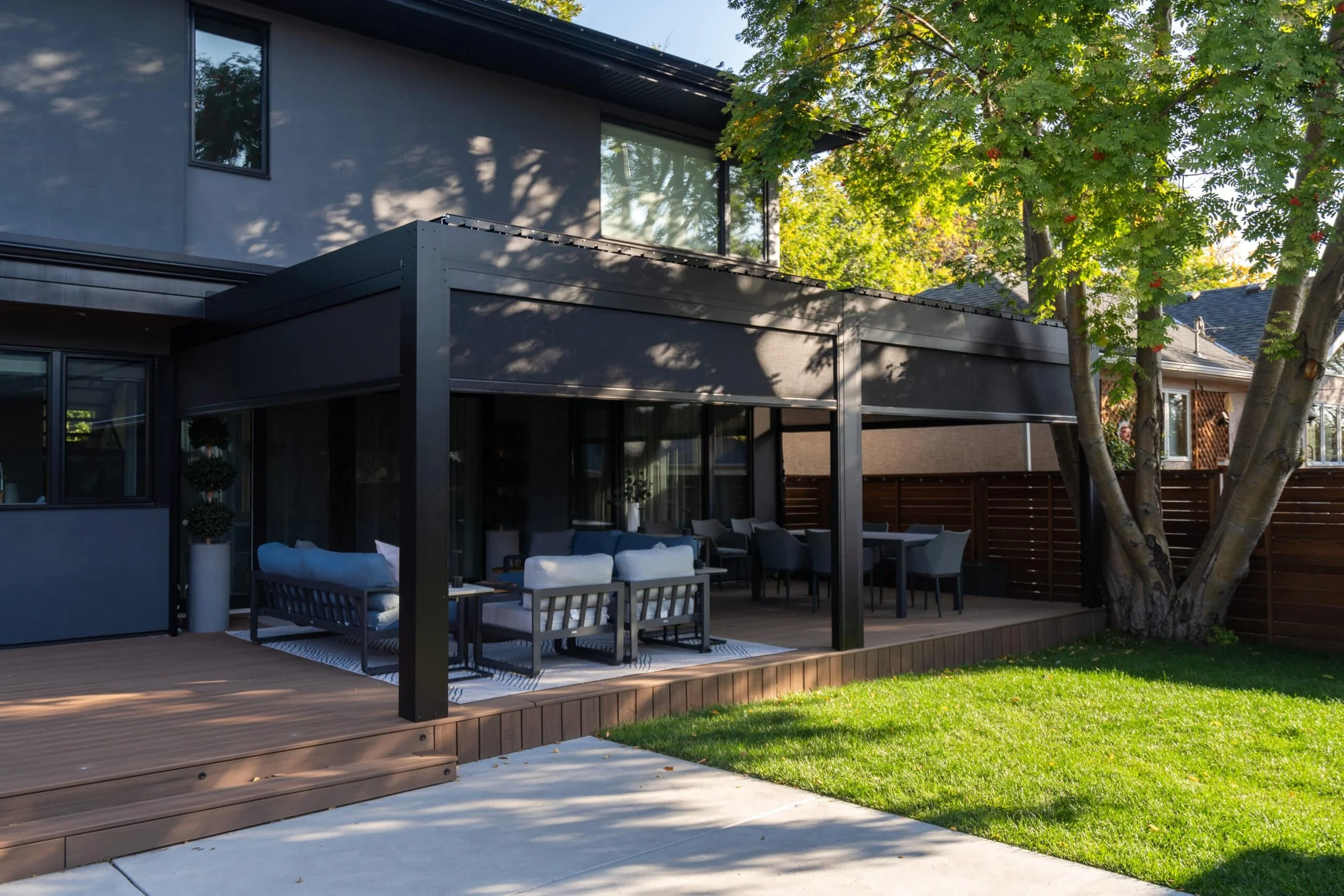 Modern house with a wooden deck patio, outdoor furniture, and a large tree with green leaves in a backyard.