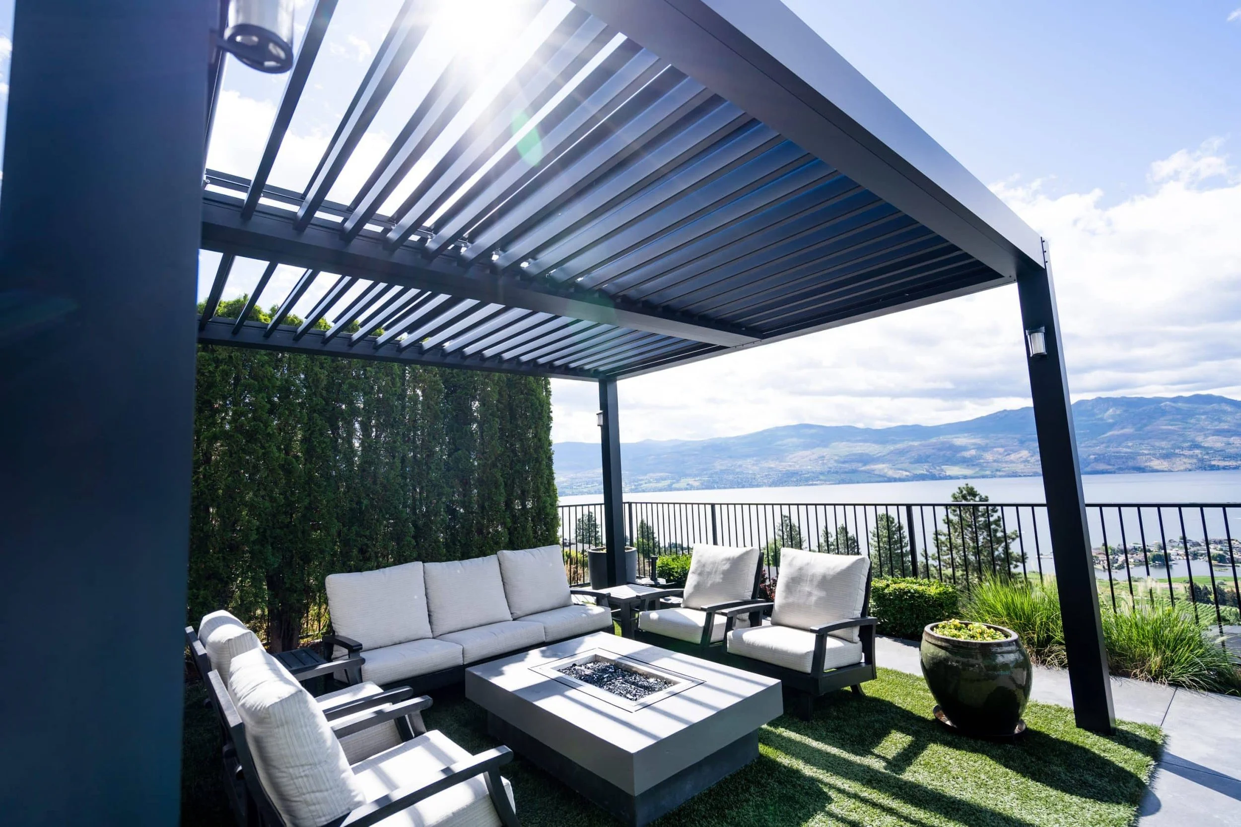 Outdoor patio with white cushioned seating under a black pergola, overlooking a scenic lake and mountains in the background, with potted plants and greenery.