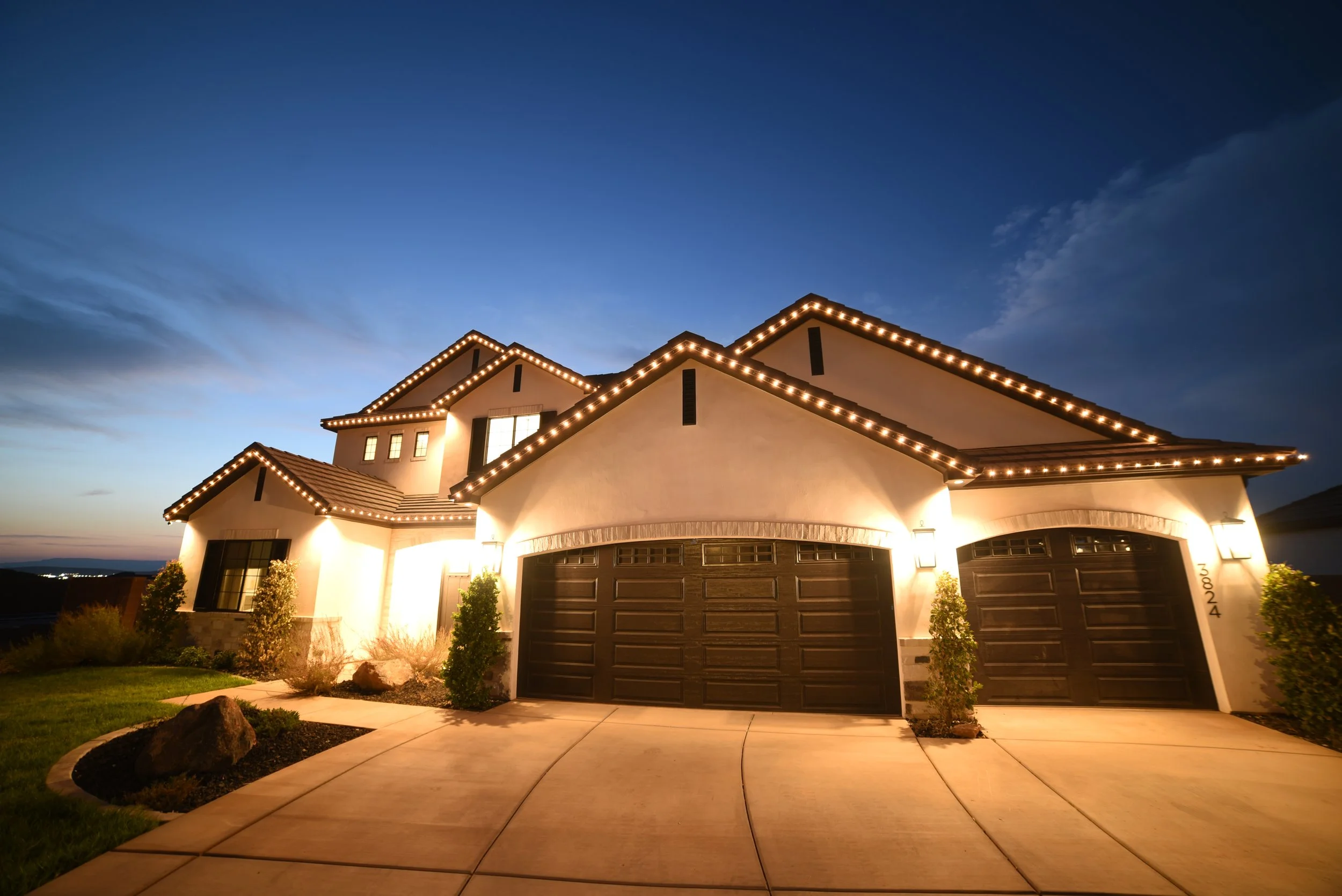 A two-story house illuminated with string lights on the roofline, featuring three garage doors and a curved driveway, during twilight.