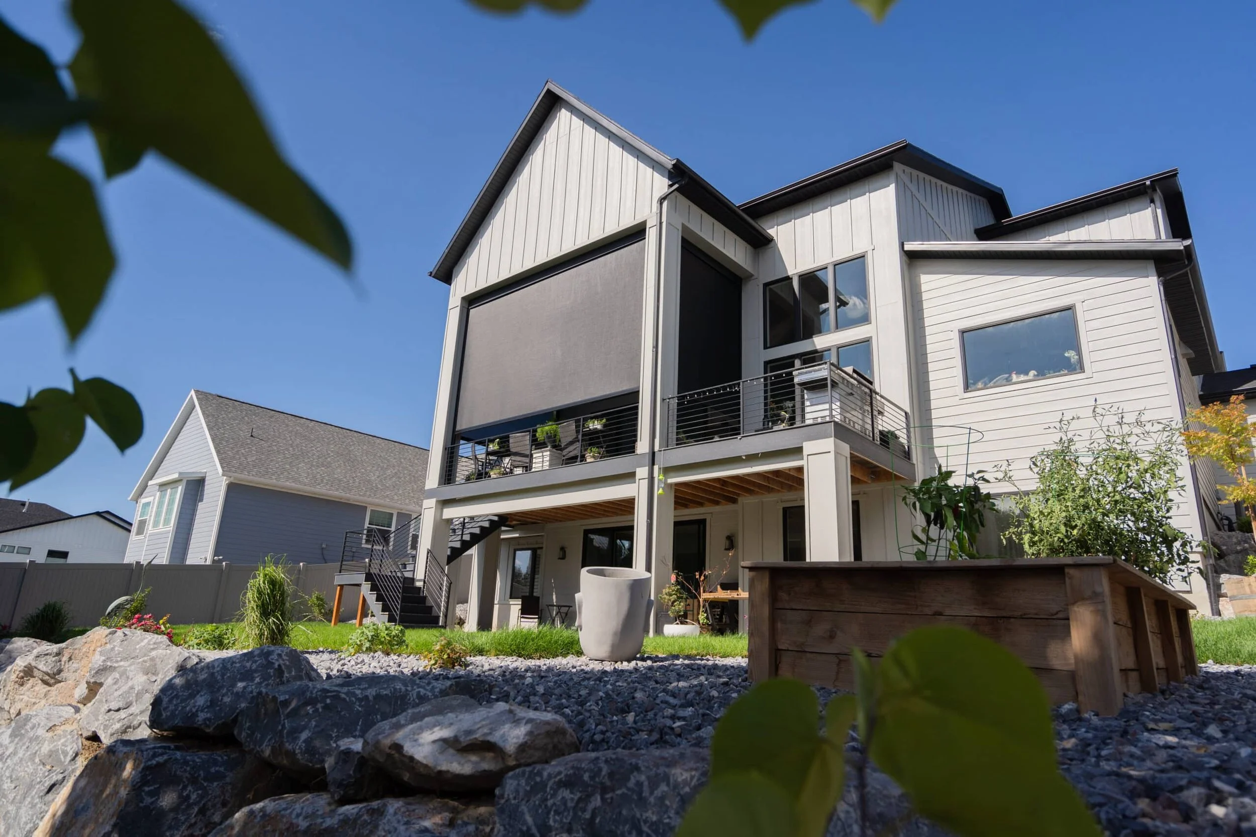 Rear view of a modern multi-story house with a balcony and outdoor staircase, surrounded by a rocky garden and plant beds, against a clear blue sky.