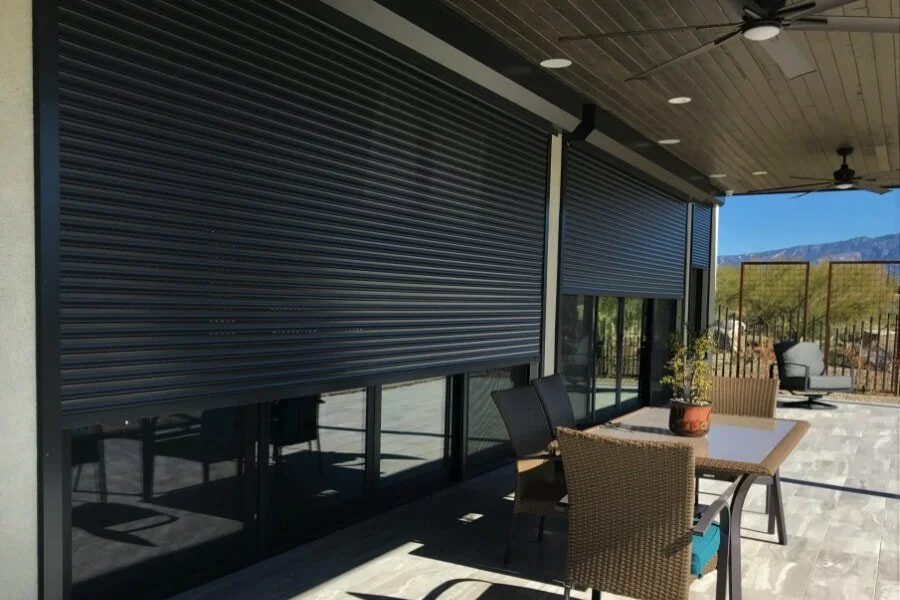 Outdoor patio with dining table, chairs, and potted plant, part of a modern house with rolling shutters and mountain view in the background.
