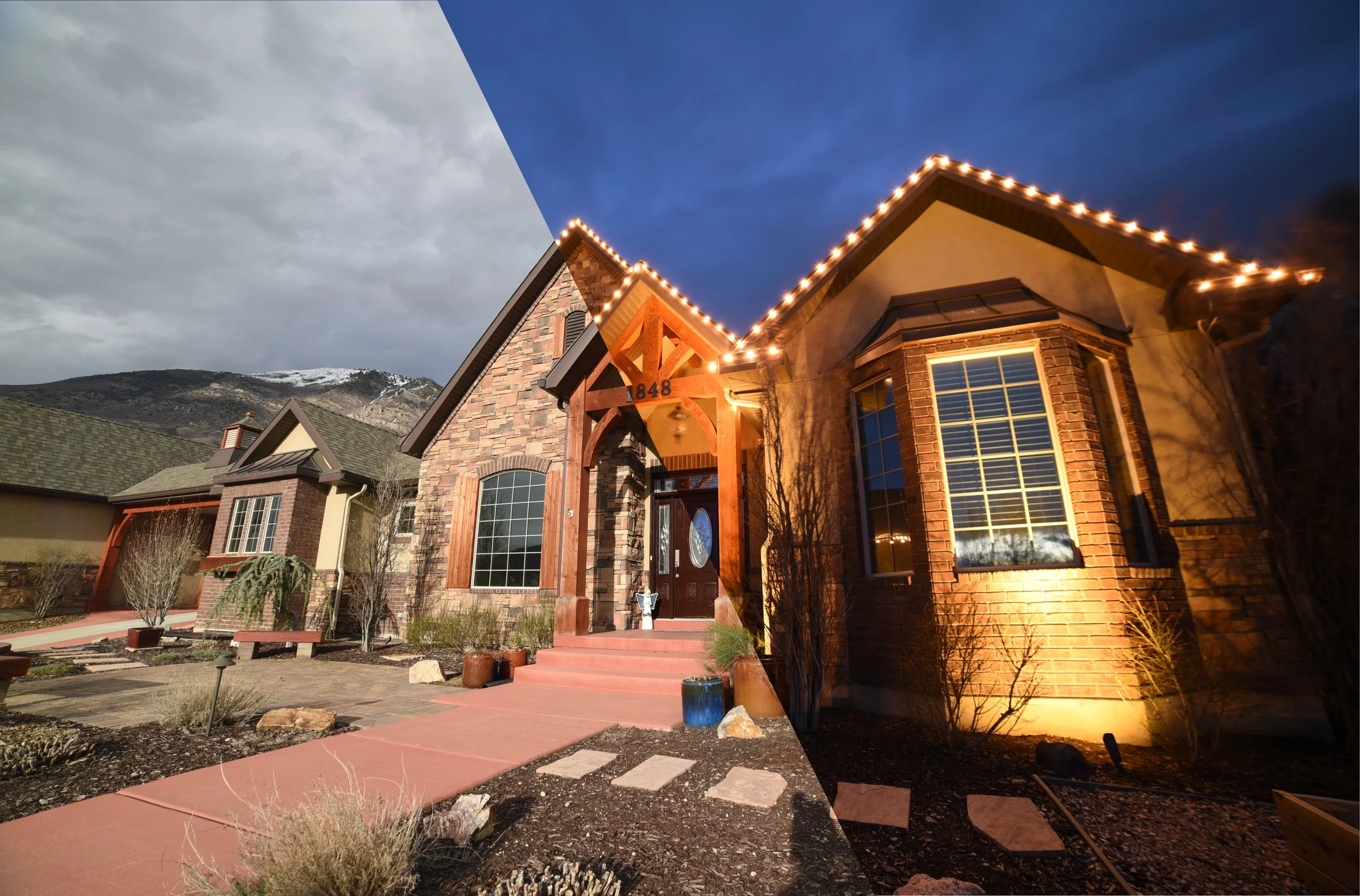 A upscale house with brick and stone exterior, illuminated with outdoor string lights at dusk or evening, snow-capped mountains in the background.