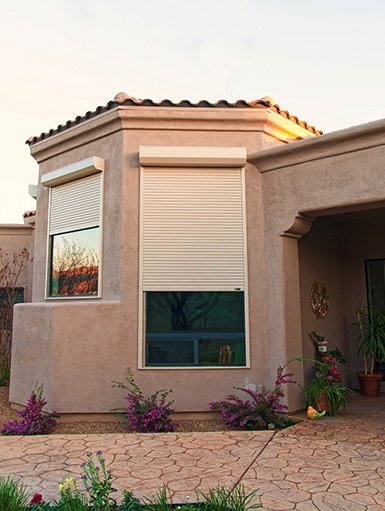 A house with beige exterior walls, two windows with white shutters, and a terracotta tile roof. The yard has a paved path and some pink flowering plants.