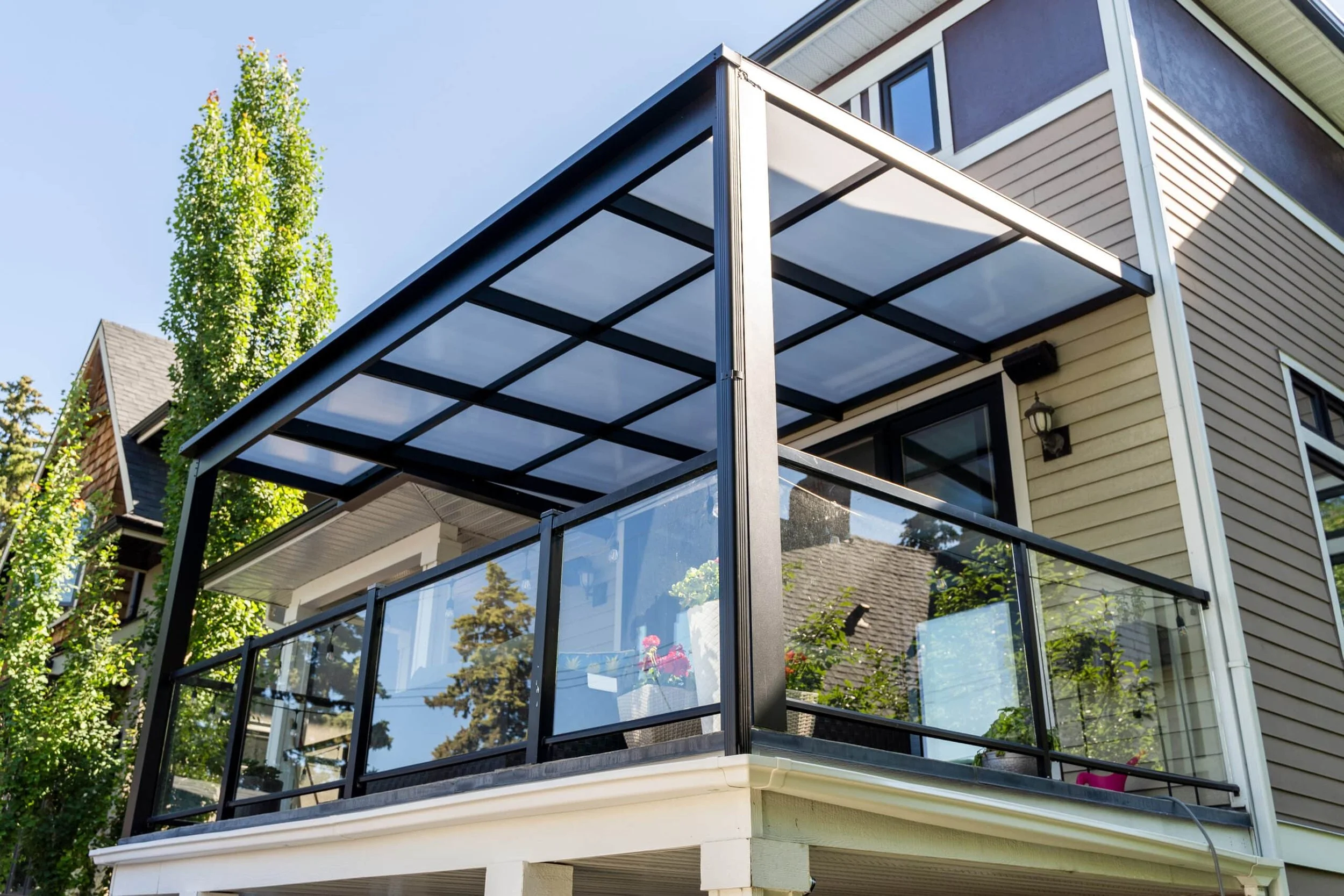 The image shows a house with a balcony or patio area featuring a black metal frame and a clear or semi-transparent roof, surrounded by a glass railing with metal support. The house has beige siding and white trim, with an exterior light fixture next to a sliding glass door leading to the balcony. The background includes blue sky and green trees.