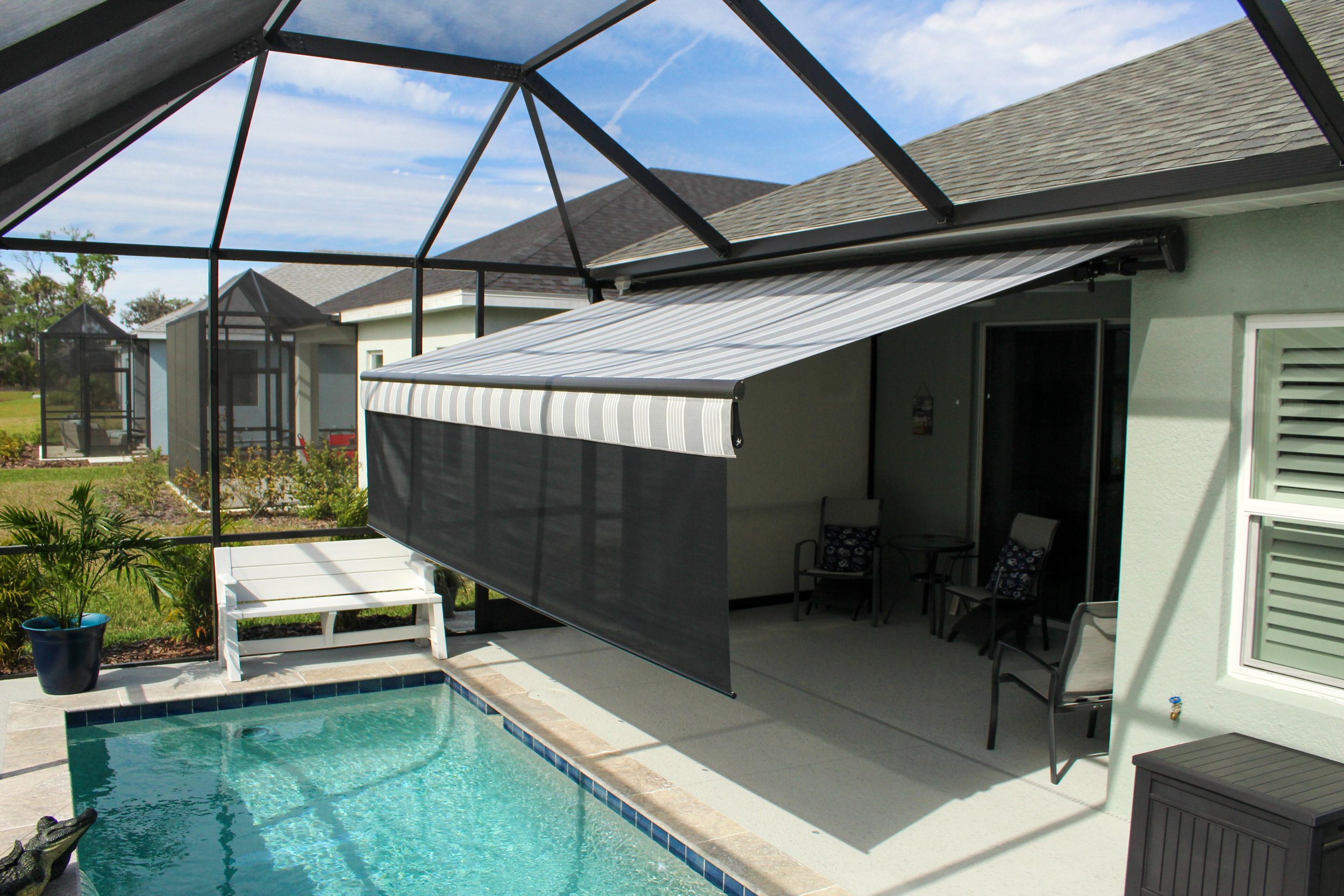 Backyard patio with a swimming pool, patio chairs, potted plants, and a screened enclosure with retractable awnings on a sunny day.