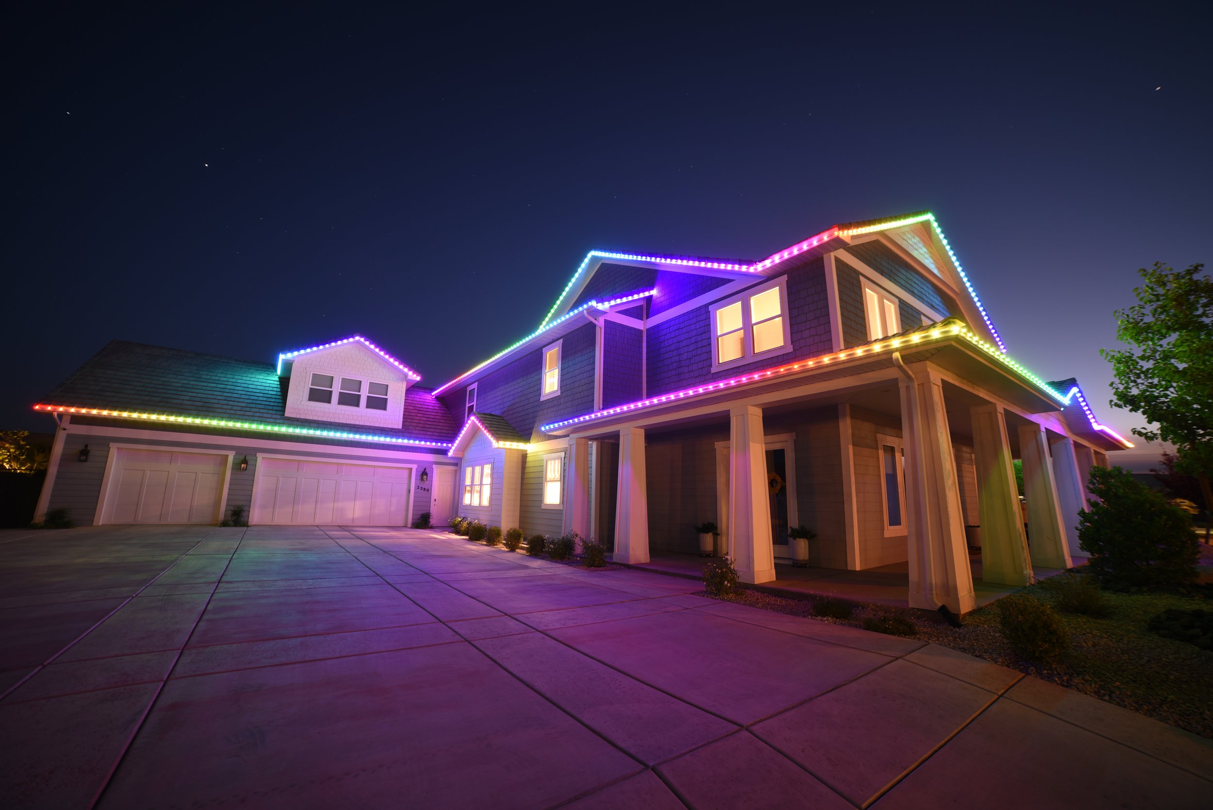 A large two-story house decorated with colorful rainbow-themed Christmas lights at night, with an empty driveway in the foreground.