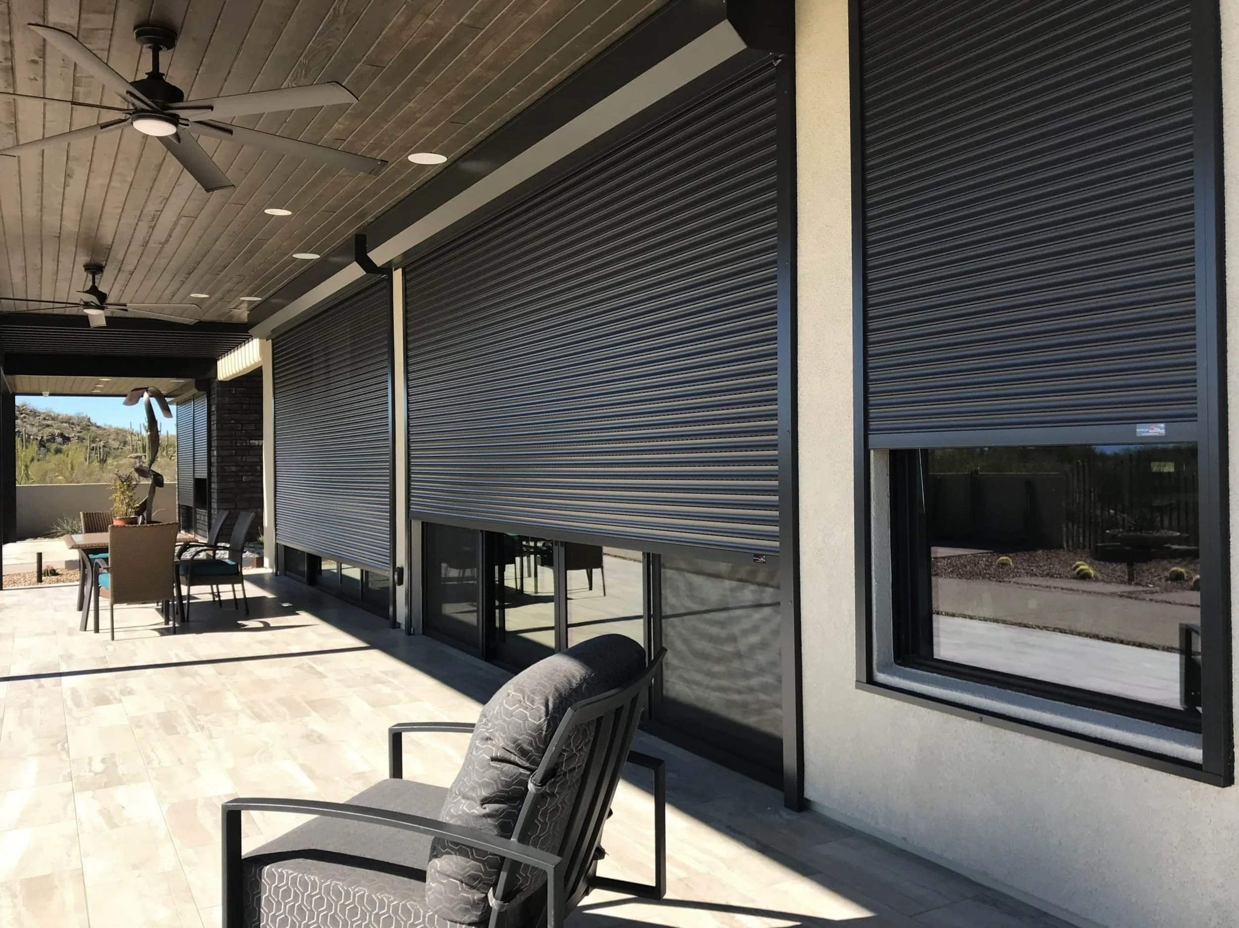Modern patio with outdoor seating, black roller shutters on house windows, ceiling fans, and a scenic background.