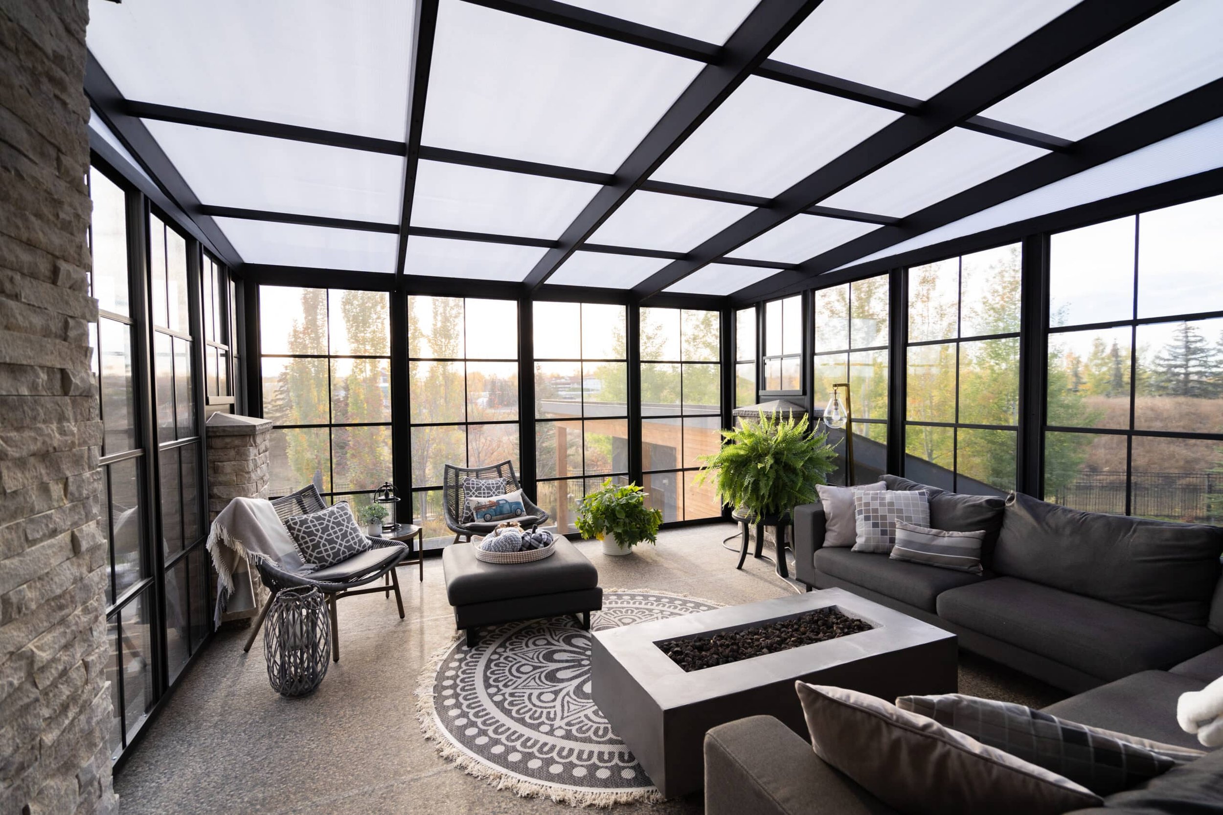 Interior view of a screened porch with large windows, a black metal ceiling, and outdoor furniture including a gray sectional sofa, chairs, and a coffee table with a fire pit insert. There are potted plants and a patterned rug on the floor.