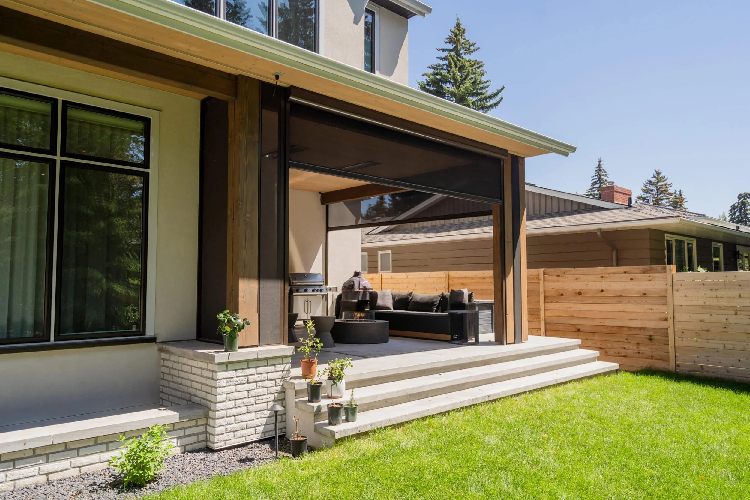 Backyard patio with outdoor furniture, potted plants, and a grill, adjacent to a house with large windows and a wood-fenced yard under a clear sky.