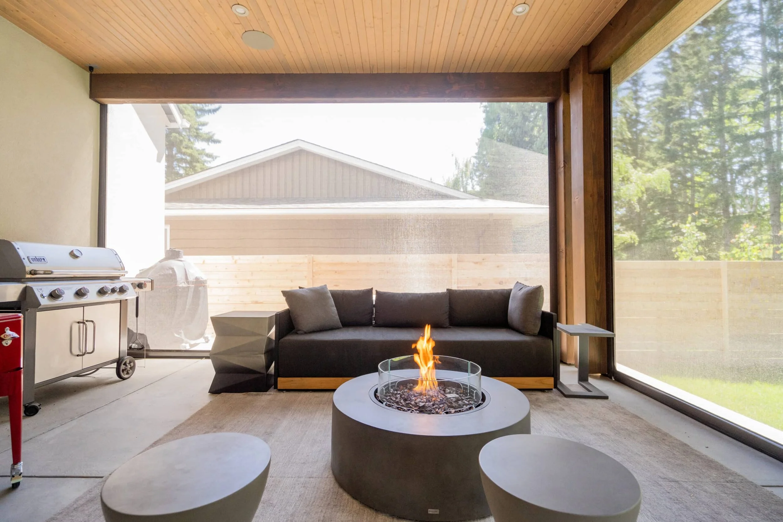 Modern living room with a black couch, fire pit table, large windows showing trees outside, and a grill on the left.