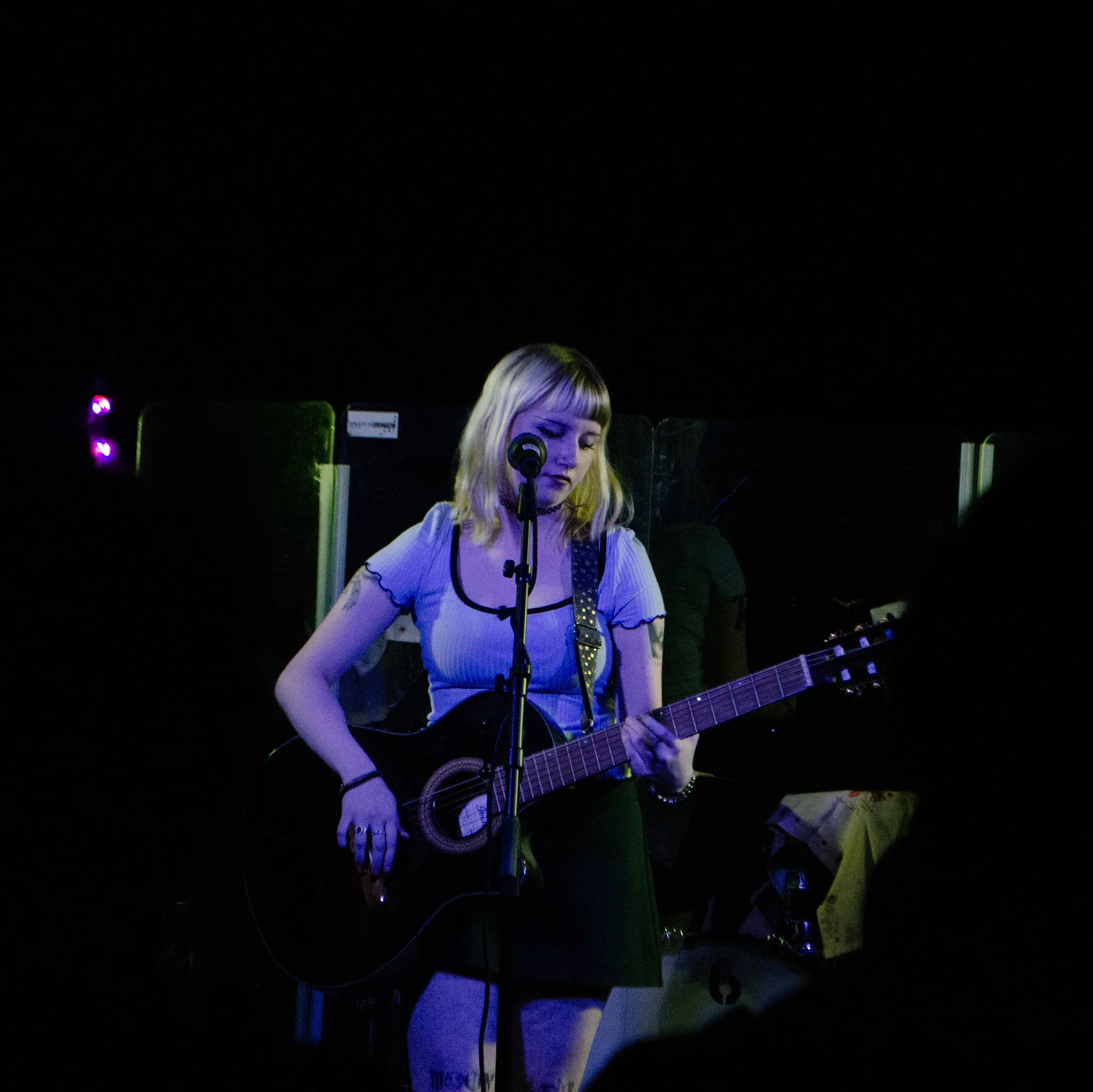 Lucretia Strokes playingacoustic guitar on stage, standing in front of a microphone, with dark background and colorful stage lighting.