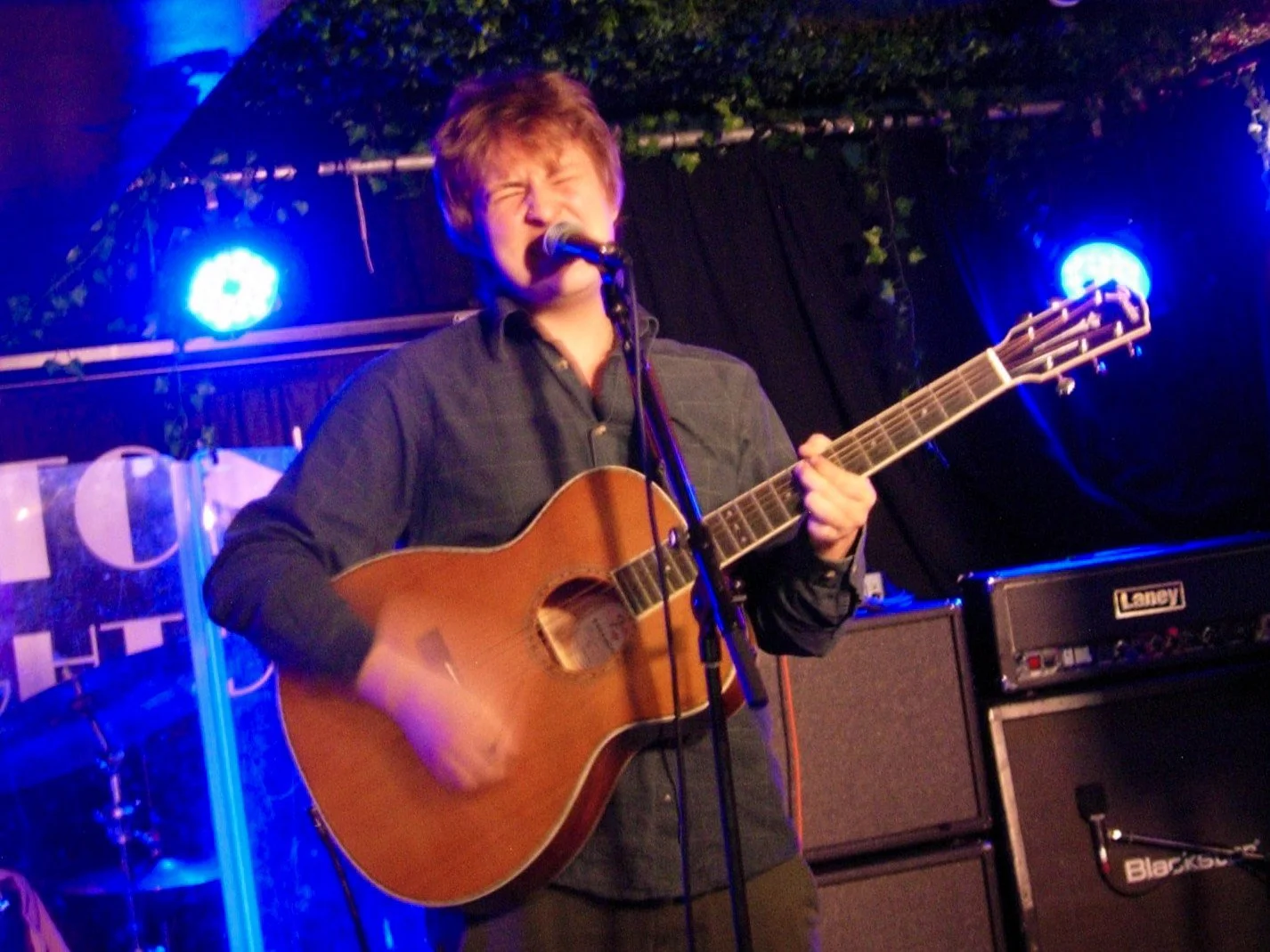 Fordy singing and playing an acoustic guitar on stage with blue stage lights and musical equipment behind him.