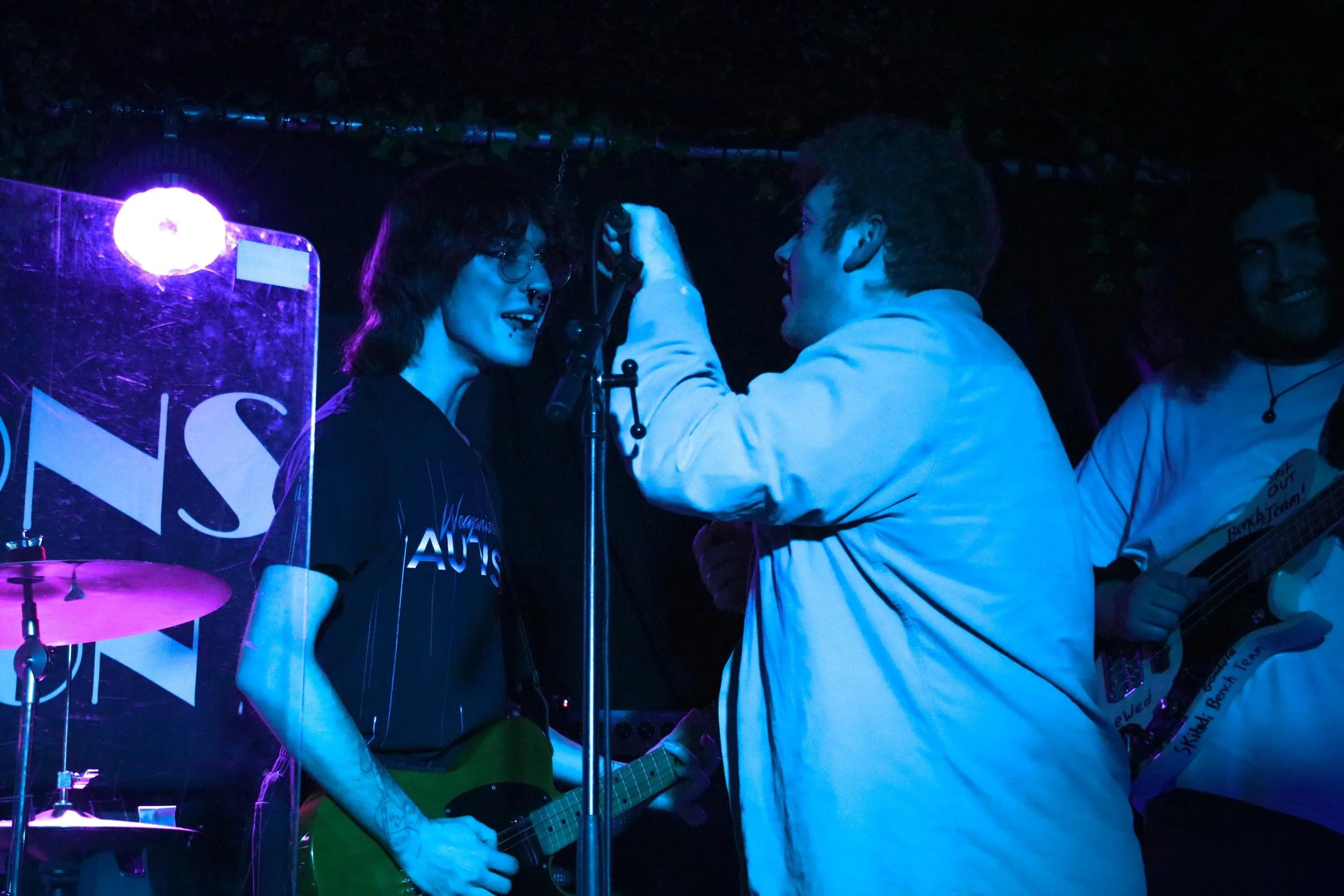 Two people on stage, one playing a yellow electric guitar and the other adjusting a microphone, with a third person smiling in the background, illuminated by blue and purple stage lights.