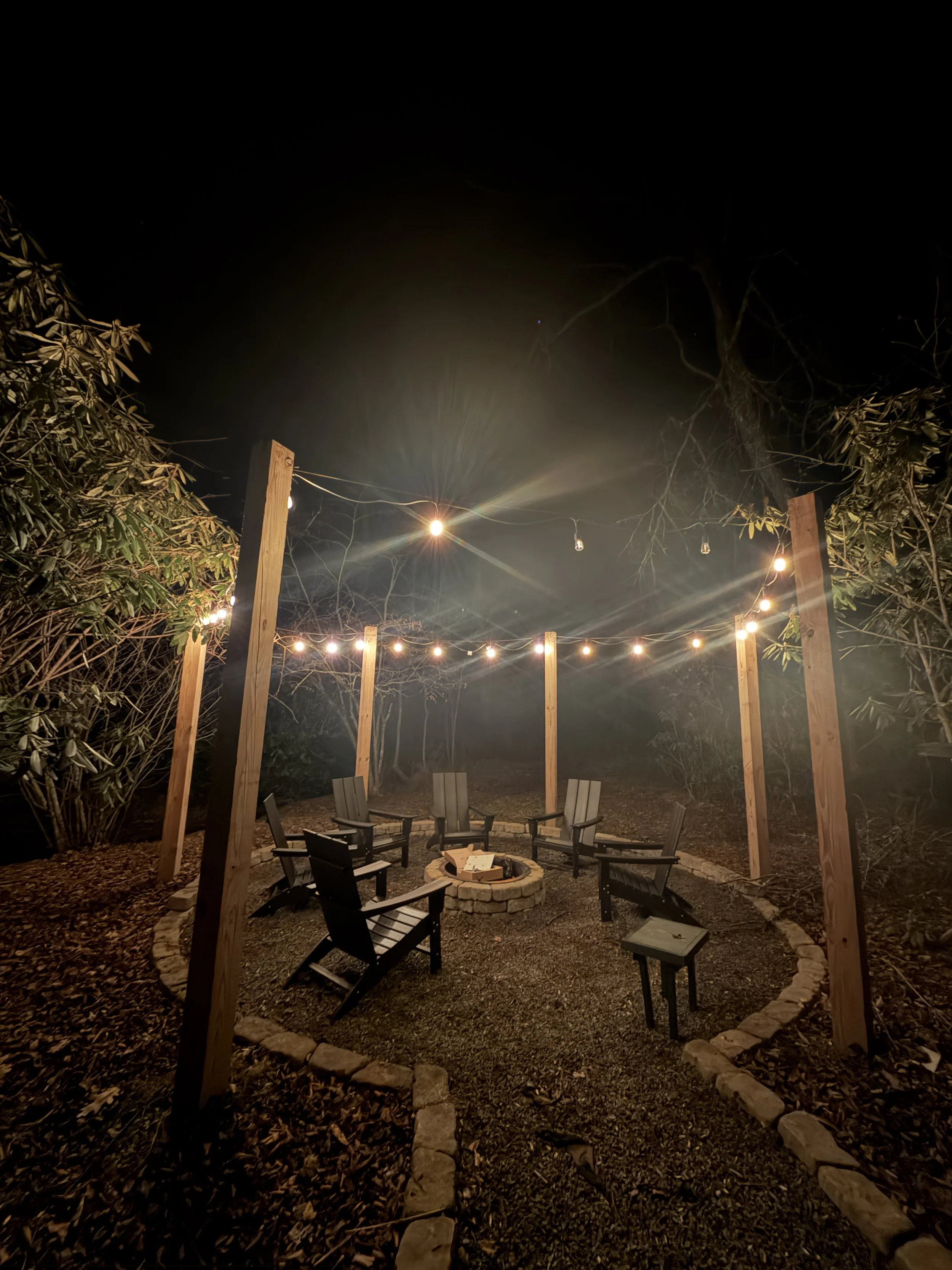 Nighttime outdoor fire pit area with six Adirondack chairs arranged in a circle around a fire pit, string lights overhead, surrounded by trees and foliage.