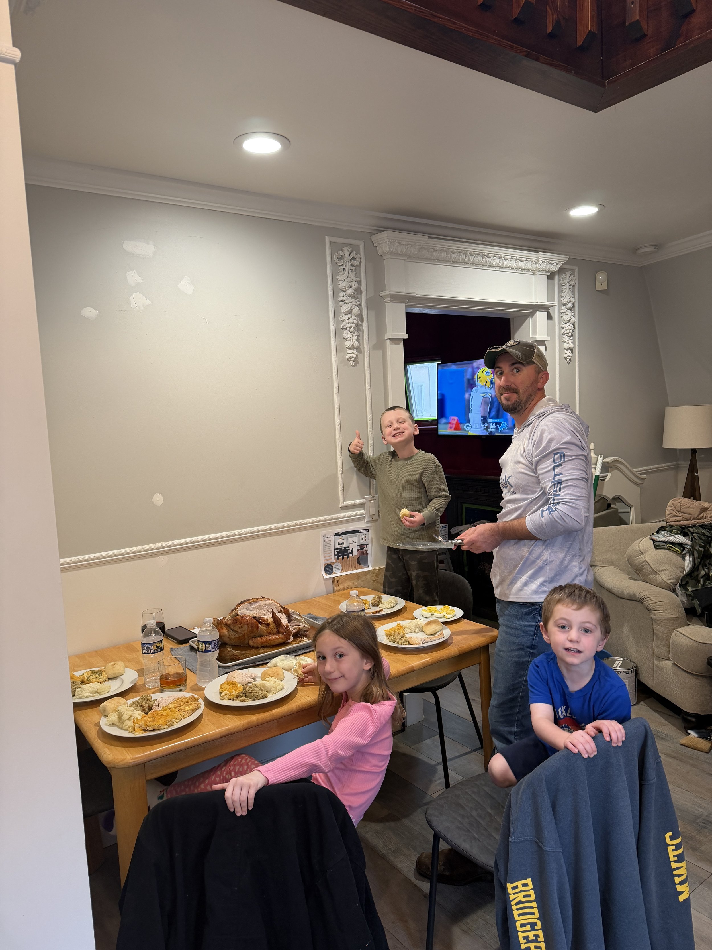 A family gathered around a dining table with a roasted turkey and various side dishes, enjoying a meal in a cozy living room with a TV and decorative white molding.