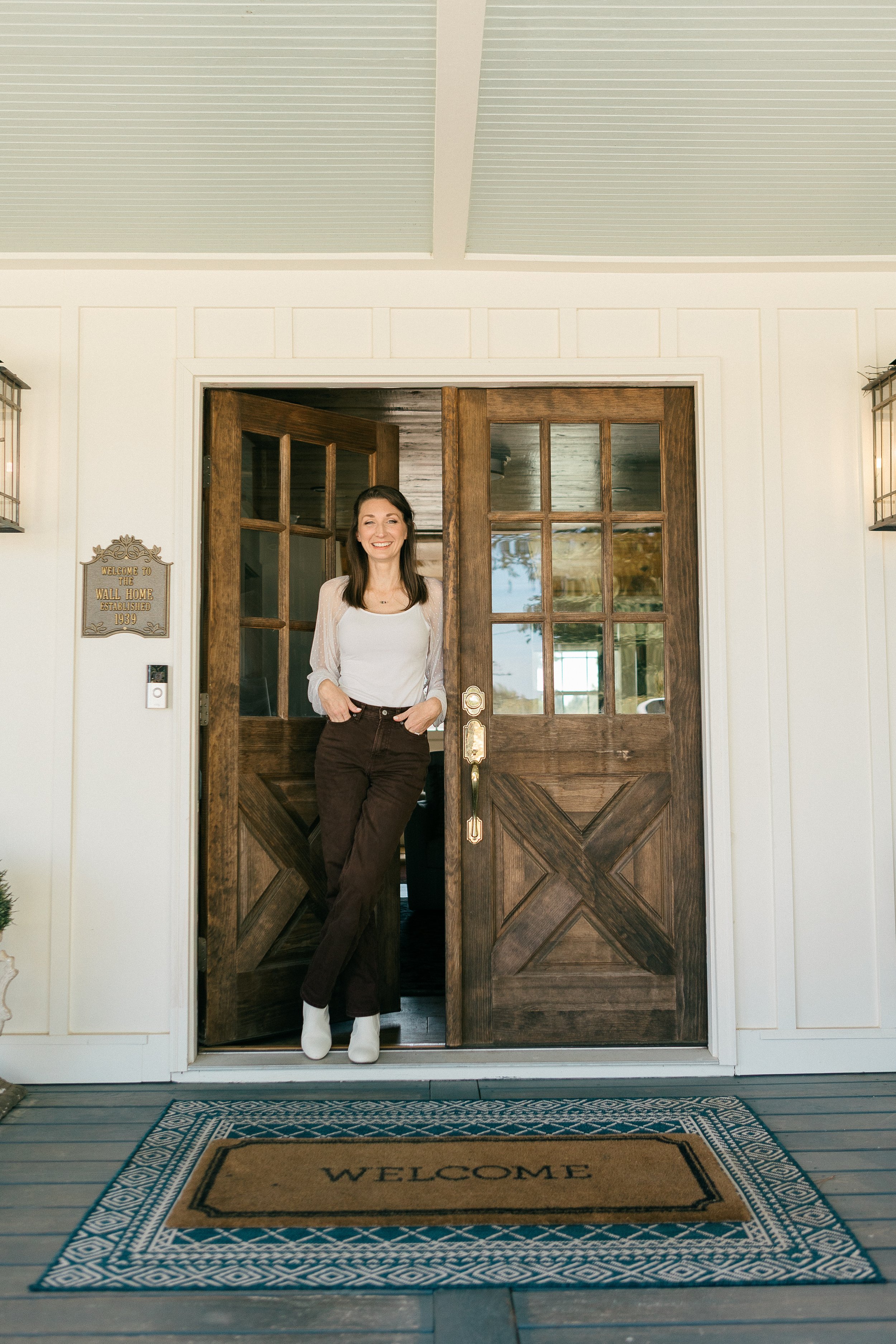Woman standing in an open wooden door, smiling, on a porch with a welcome mat that says 'WELCOME'.