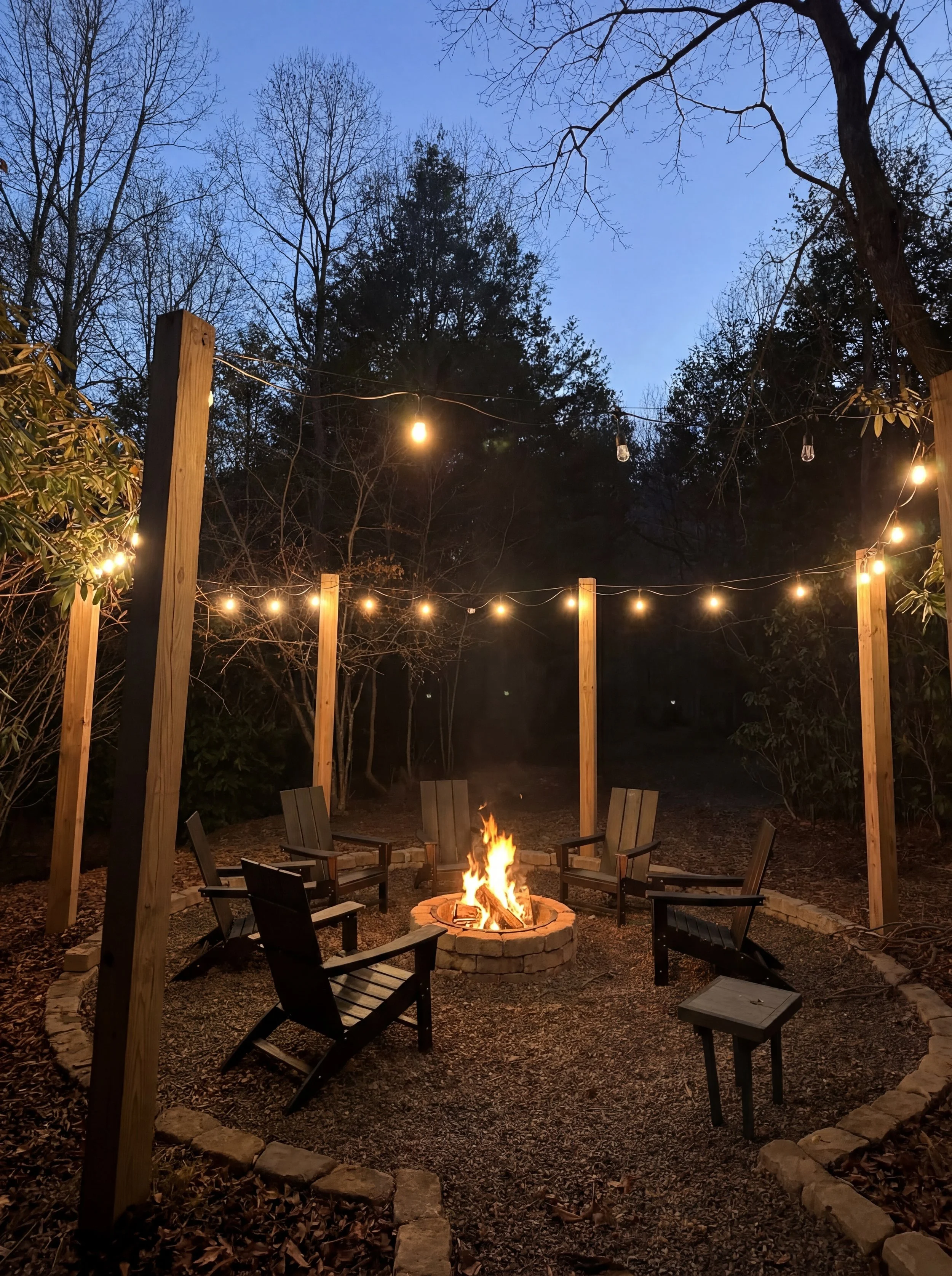 Nighttime outdoor fire pit area with six Adirondack chairs arranged in a circle around a fire pit, string lights overhead, surrounded by trees and foliage.