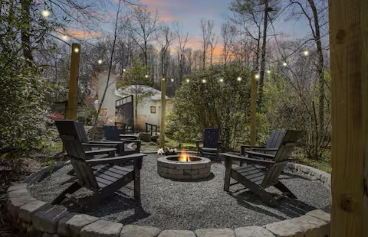 Outdoor seating area with six wooden chairs arranged in a circle around a fire pit. String lights hang above, and there are trees and a small shed in the background during dusk.