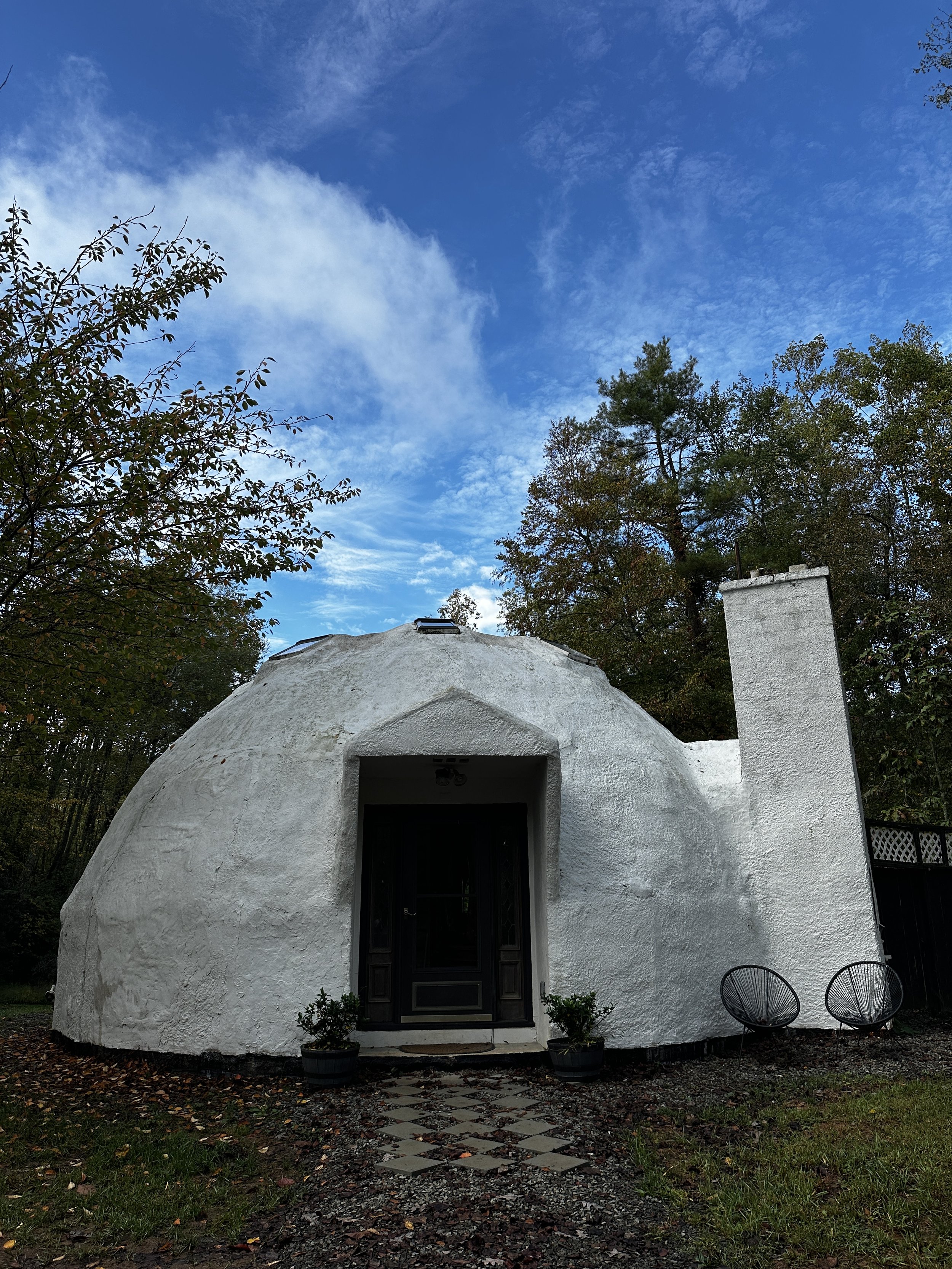 White, dome-shaped house with a black door, two potted plants at the entrance, two black round chairs outside, surrounded by trees against a blue sky with scattered clouds.