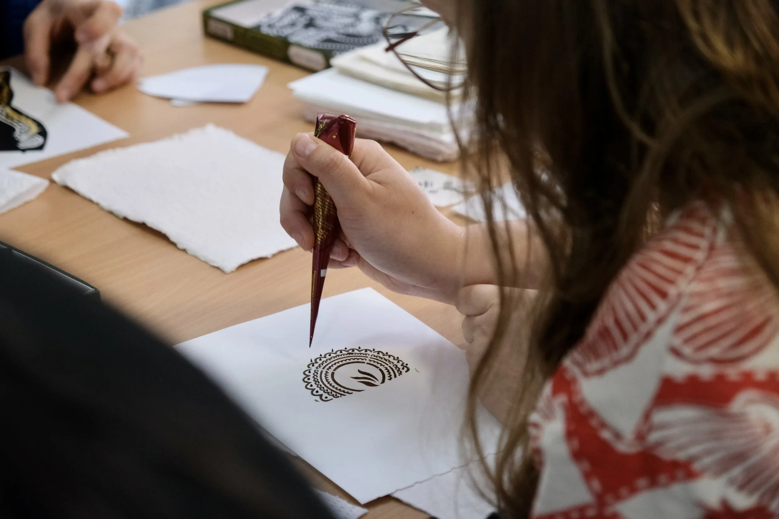 A person using a cone-shaped tool to create henna designs on a piece of white paper or fabric. The workspace has papers, a book, and tissues.