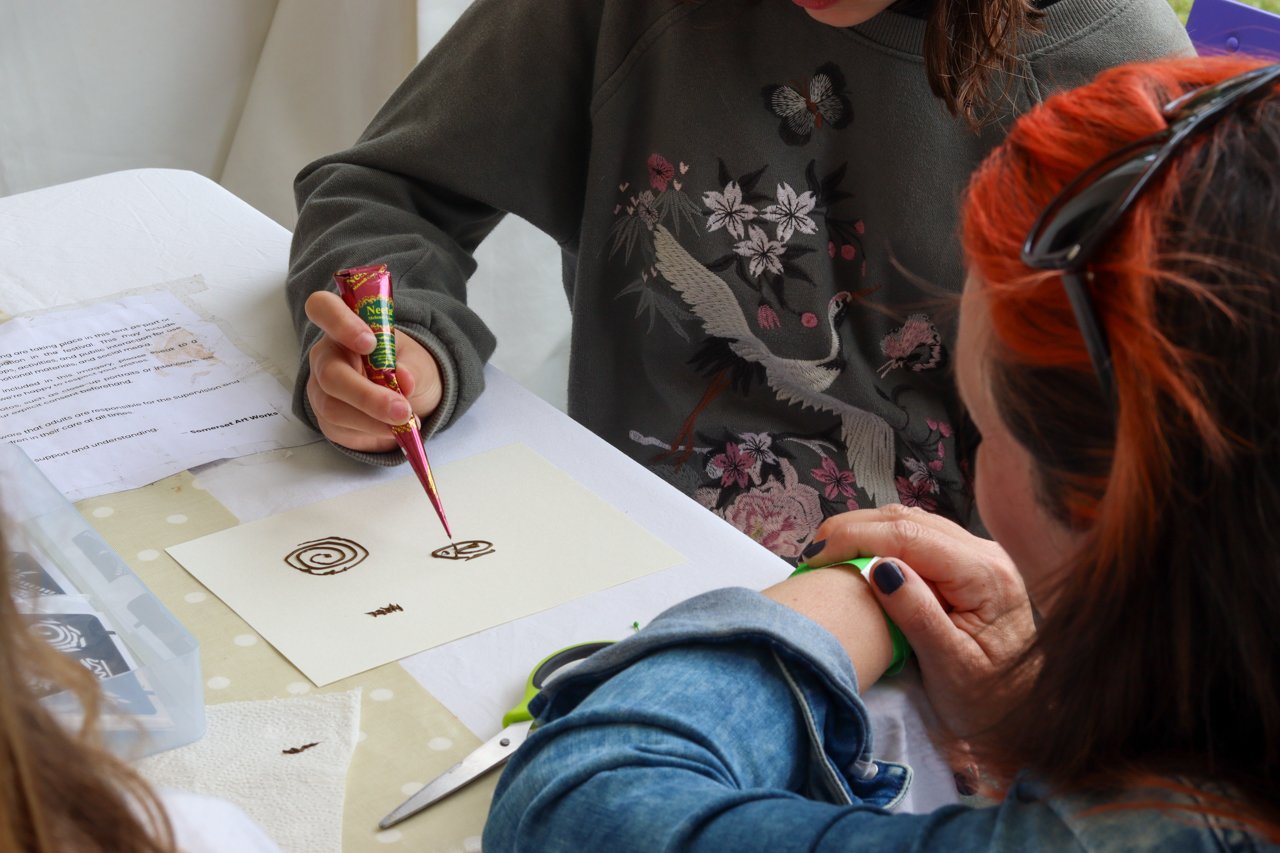 A person with red hair and sunglasses is doing a tattoo on another person's arm while sitting at a table.