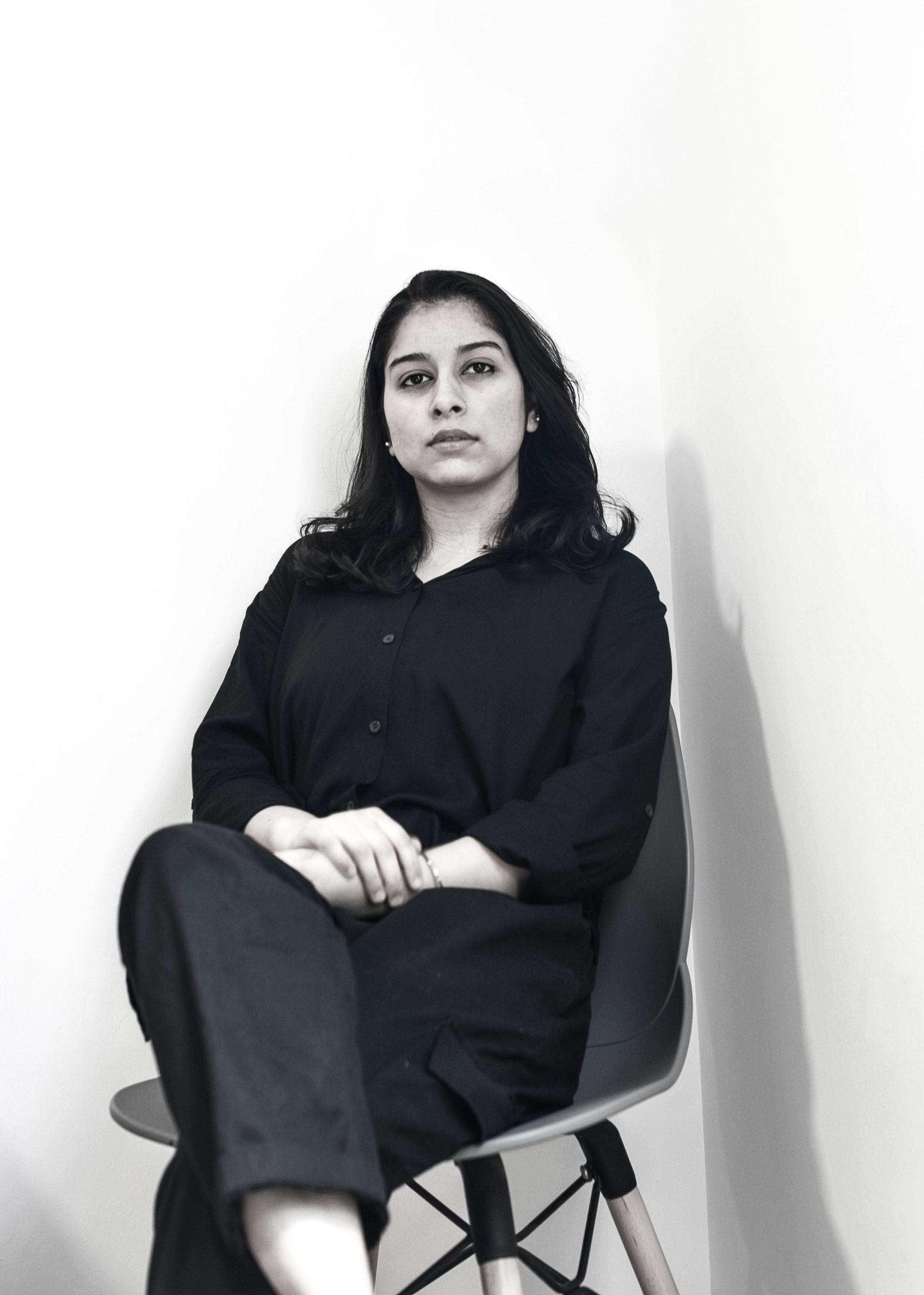 A young woman with dark, wavy hair sits on a modern, gray chair against a plain white wall. She is wearing a black button-up shirt and dark pants, looking directly at the camera with a neutral expression.