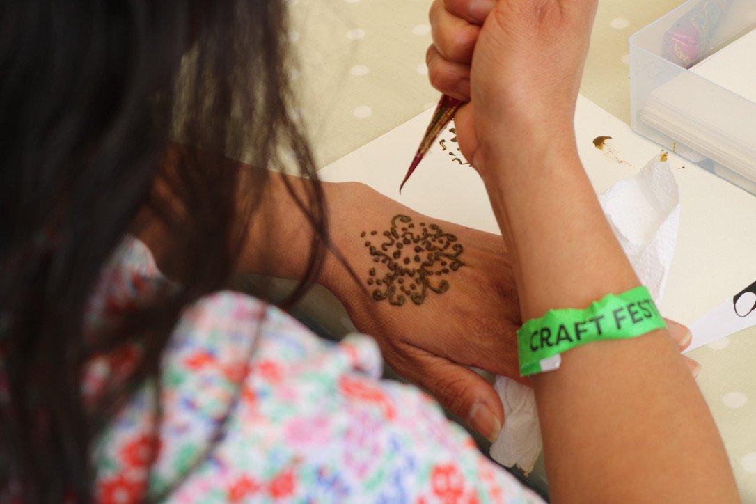 Person with dark hair and a green wristband labeled 'CRAFT FEST' is decorating a tattoo or henna design on their wrist with a small brush.