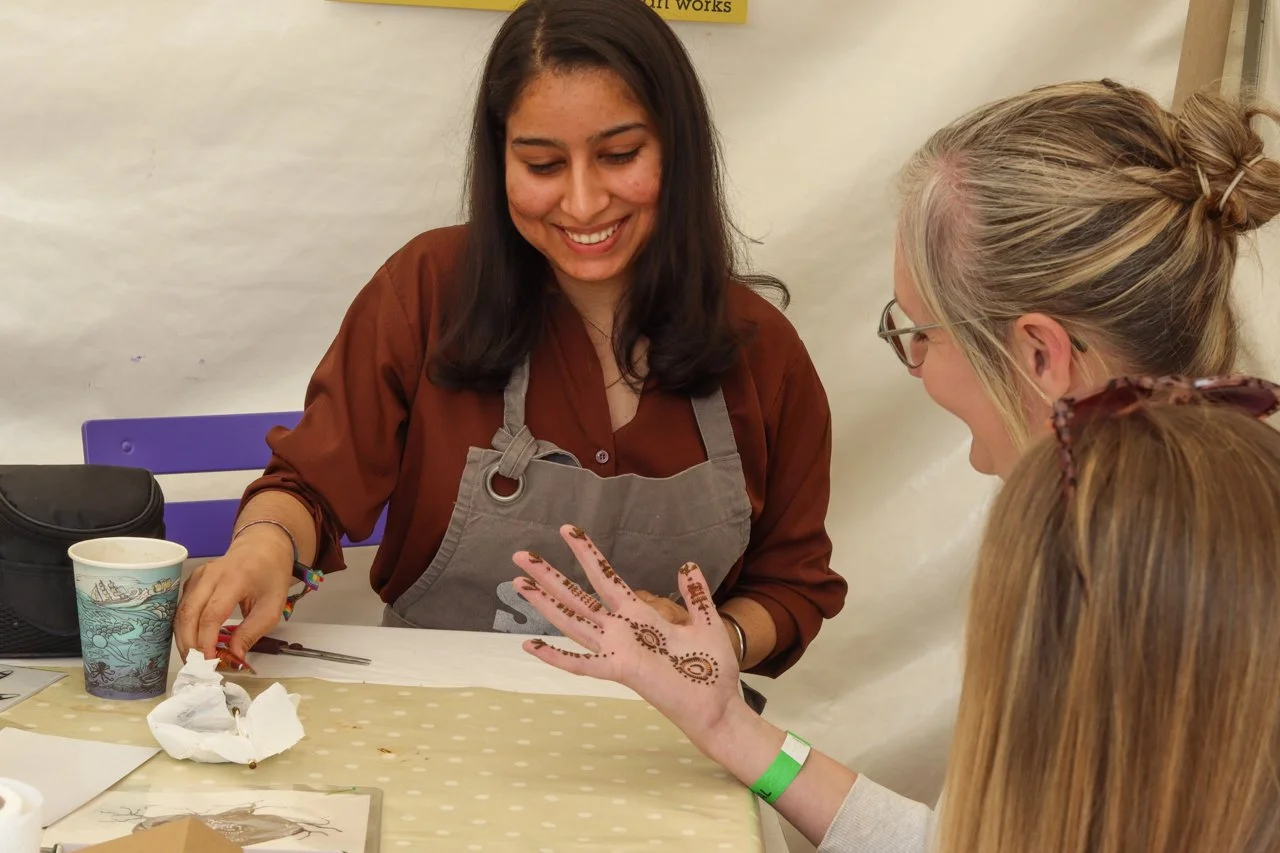 A woman with tan skin, dark hair, and a brown shirt smiles as she shows henna tattoos on her hand to two women with blonde hair. One of the blonde women has henna on her hand and wears a green wristband, and the other is seated with her head turned away.