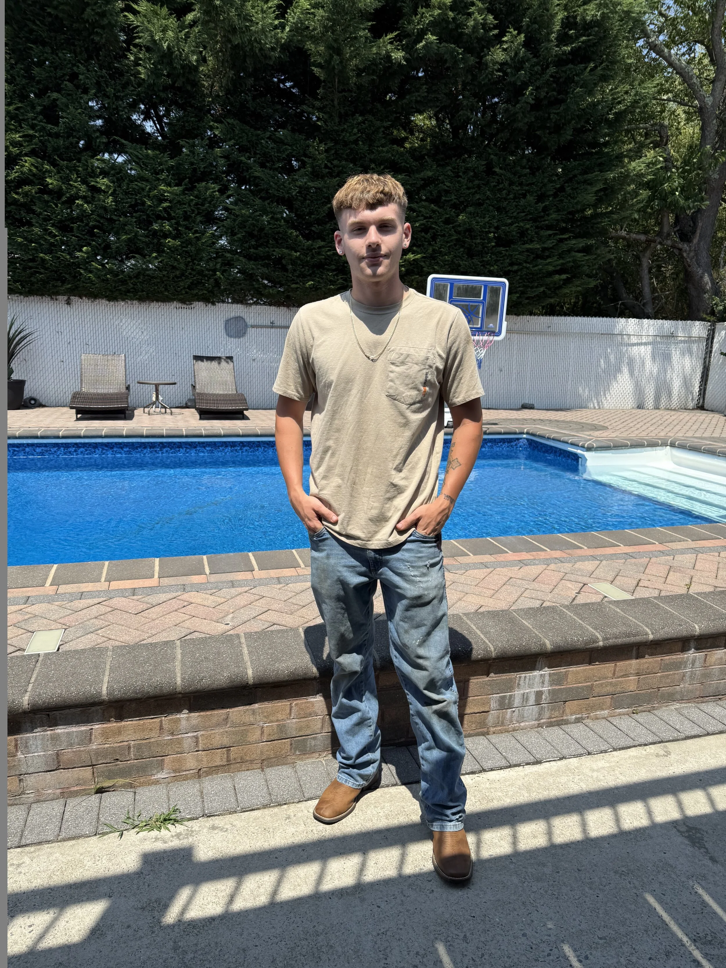 A young man standing near a backyard swimming pool with a basketball hoop in the background, wearing a beige t-shirt, blue jeans, and brown shoes.