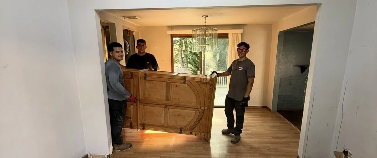 Three workers are moving a large wooden door through a doorway into a living room with a chandelier and large windows showing greenery outside.