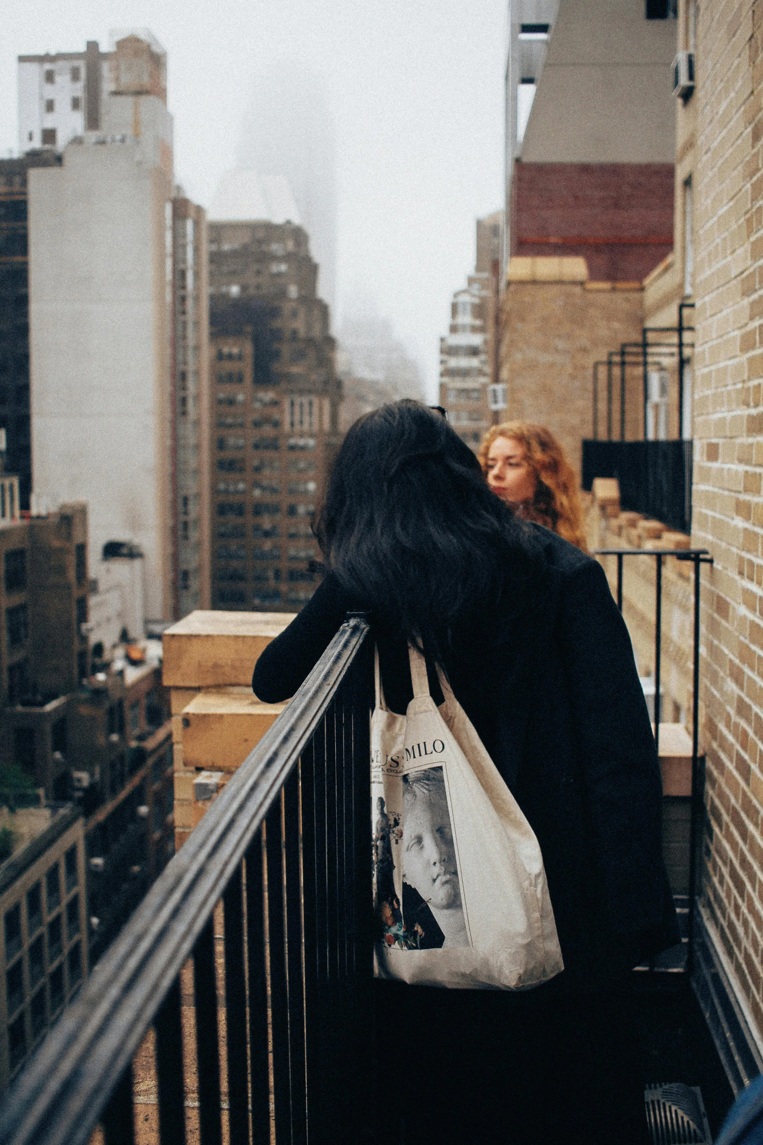 Two women on a city balcony, one with black hair leaning on the railing, the other with curly red hair looking away, with tall buildings and fog in the background.