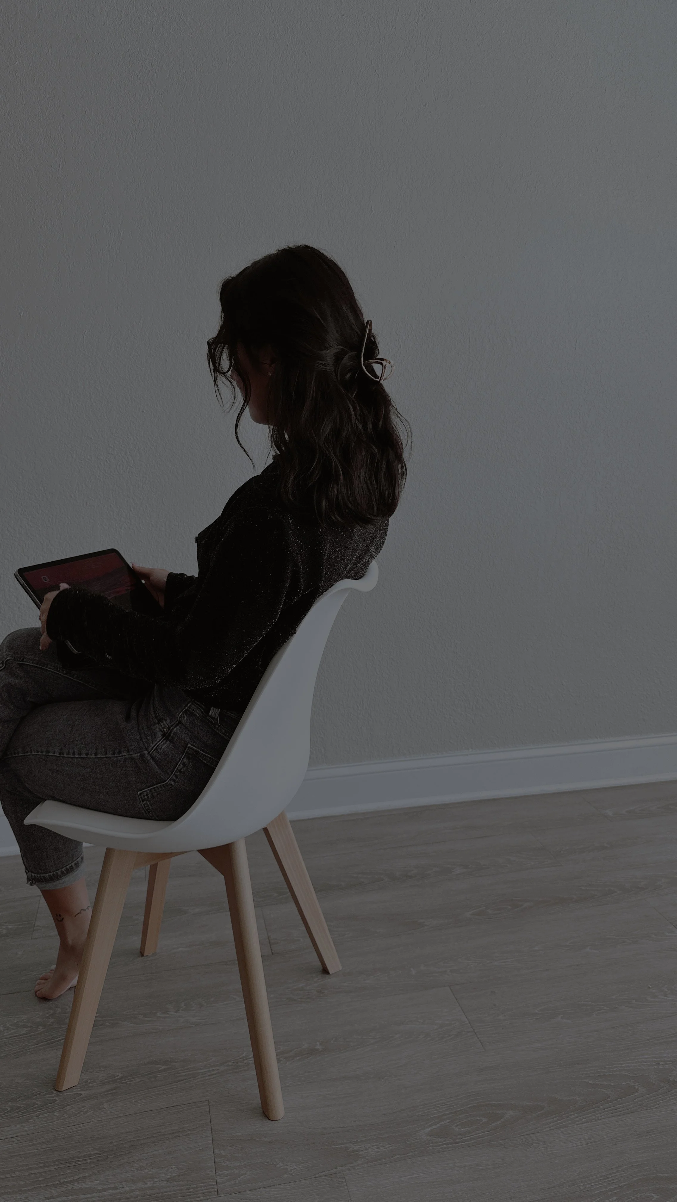 A woman with dark wavy hair sitting in a white chair with wooden legs, looking at a tablet, positioned against a plain grey wall, standing on light-colored wood flooring.