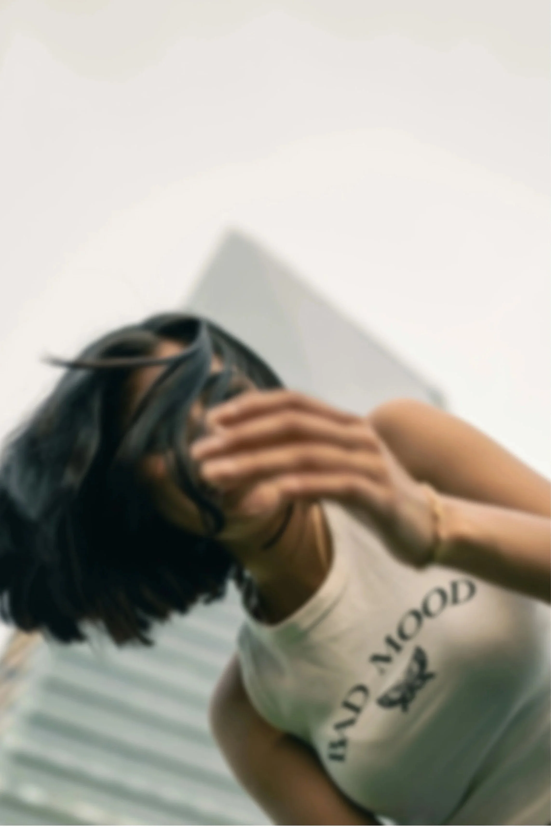 A woman with black hair and glasses pulling her hair back, wearing a sleeveless shirt with text and an illustration, standing in front of a low-angle background with white sky and a building.