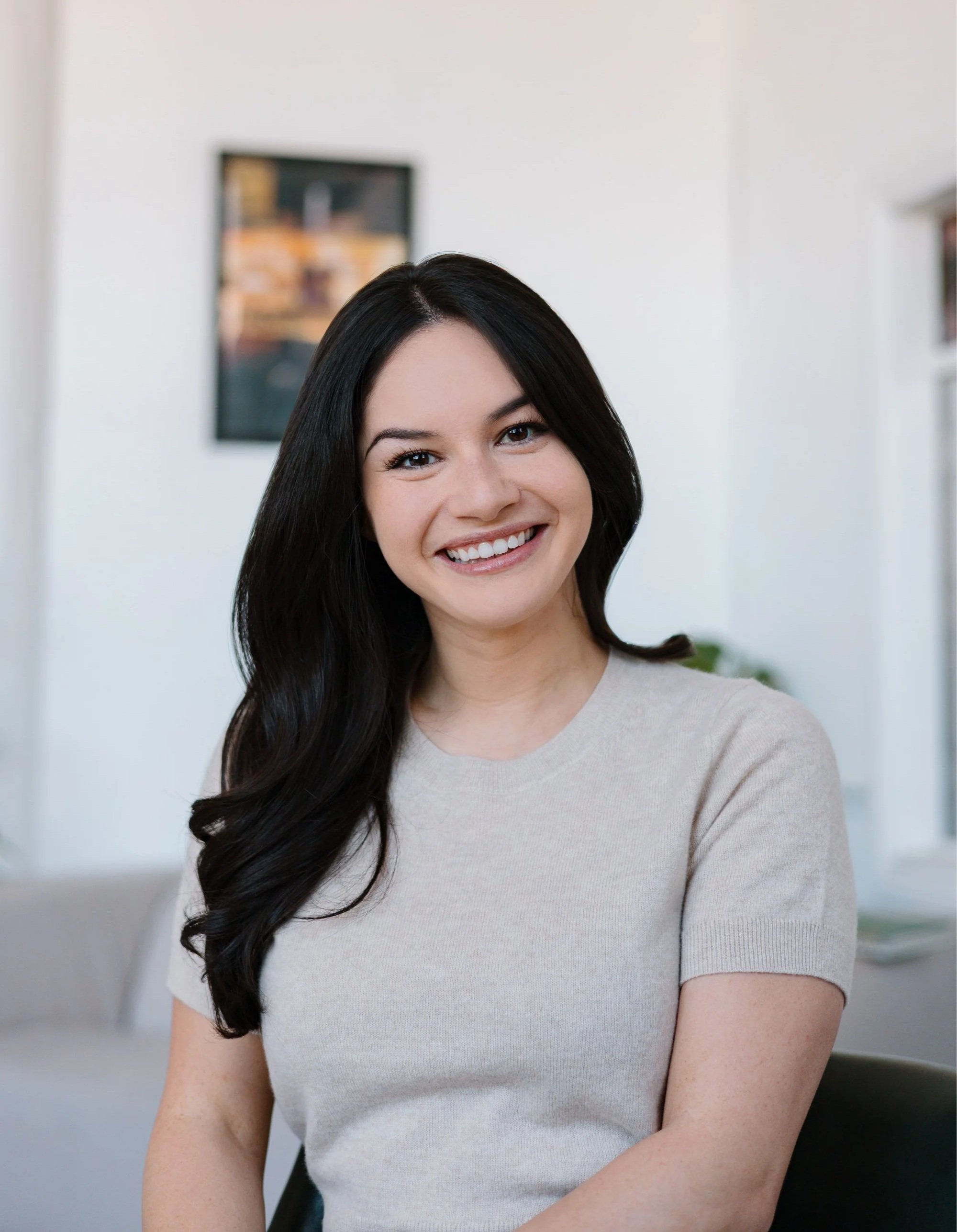 Smiling woman with long dark hair in a beige top inside a bright room.