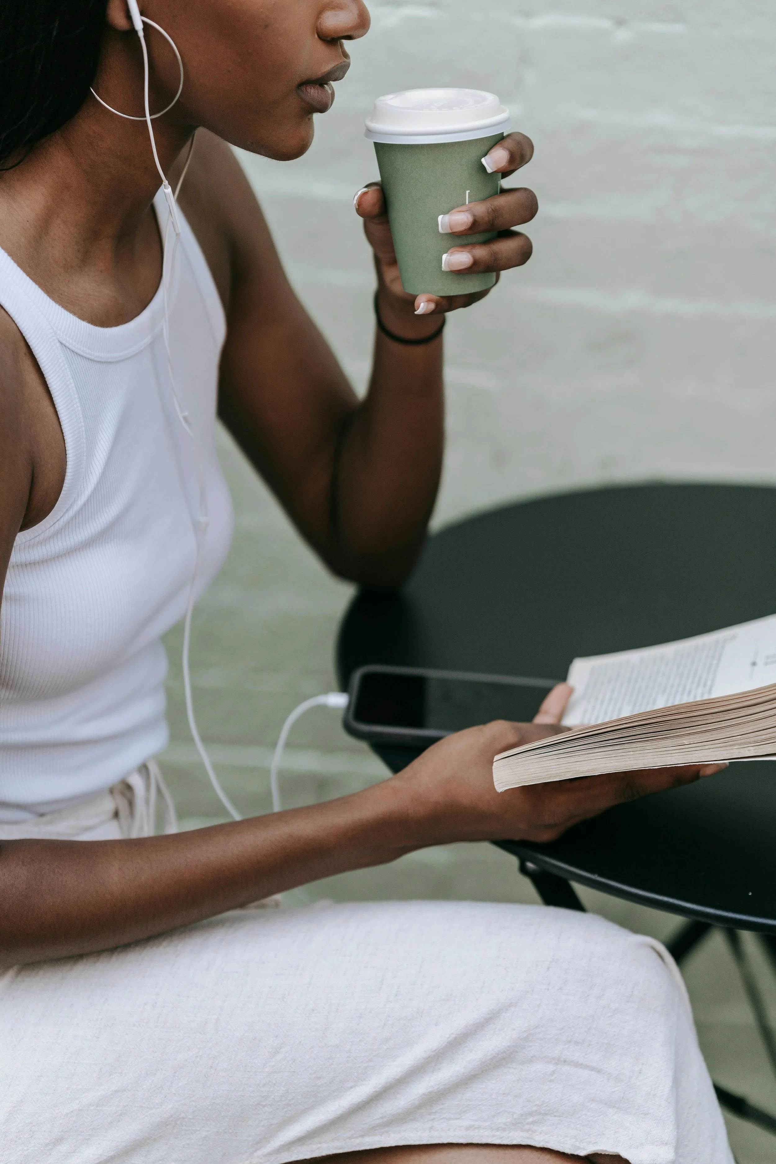 A woman with dark skin is sitting at a black table, reading a book, listening to something on her earbuds, and holding a green coffee cup.