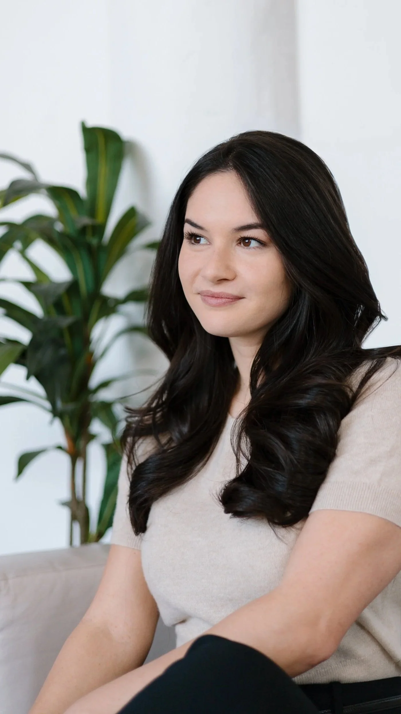 A woman with long dark hair sitting on a beige couch in a light-colored room, with a tall green plant in the background.