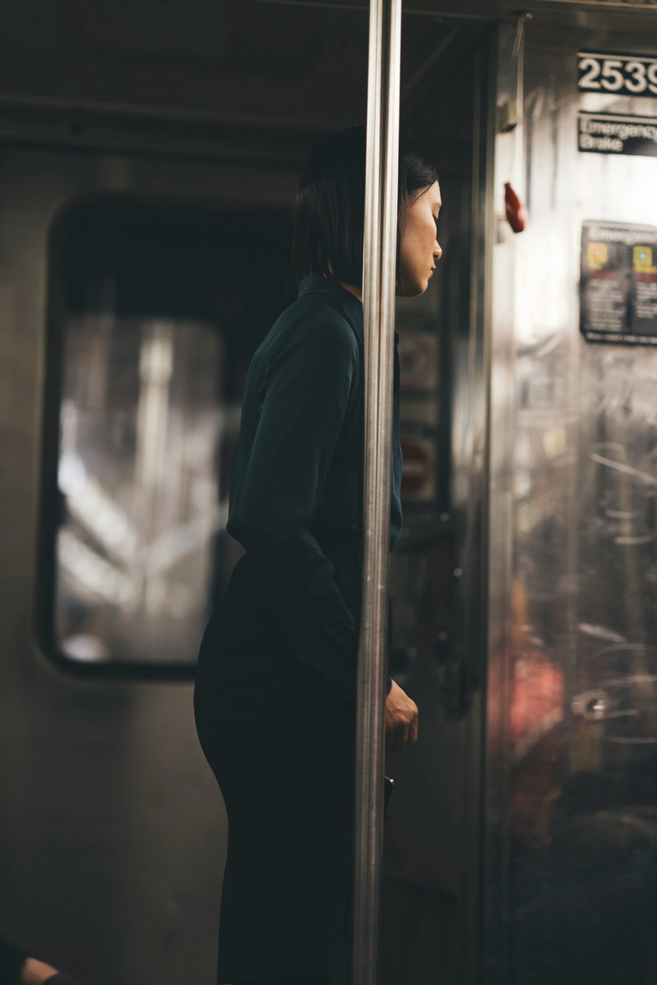 A woman standing on a subway train, seen in profile from the side, with a metallic panel obscuring half of her face and body.