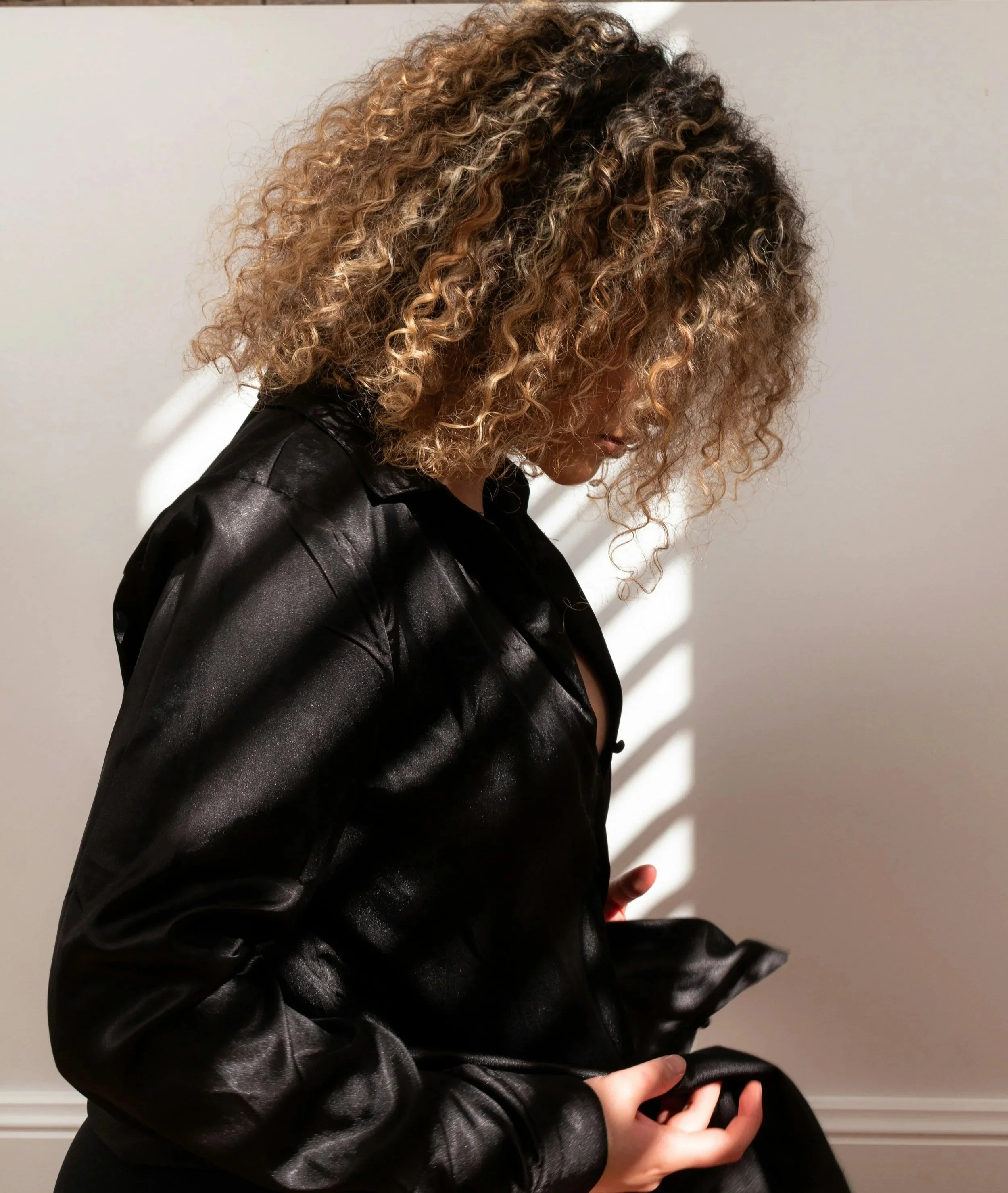 A woman with curly blonde hair looking down while holding a black satin jacket, standing against a plain light-colored wall with shadow stripes.