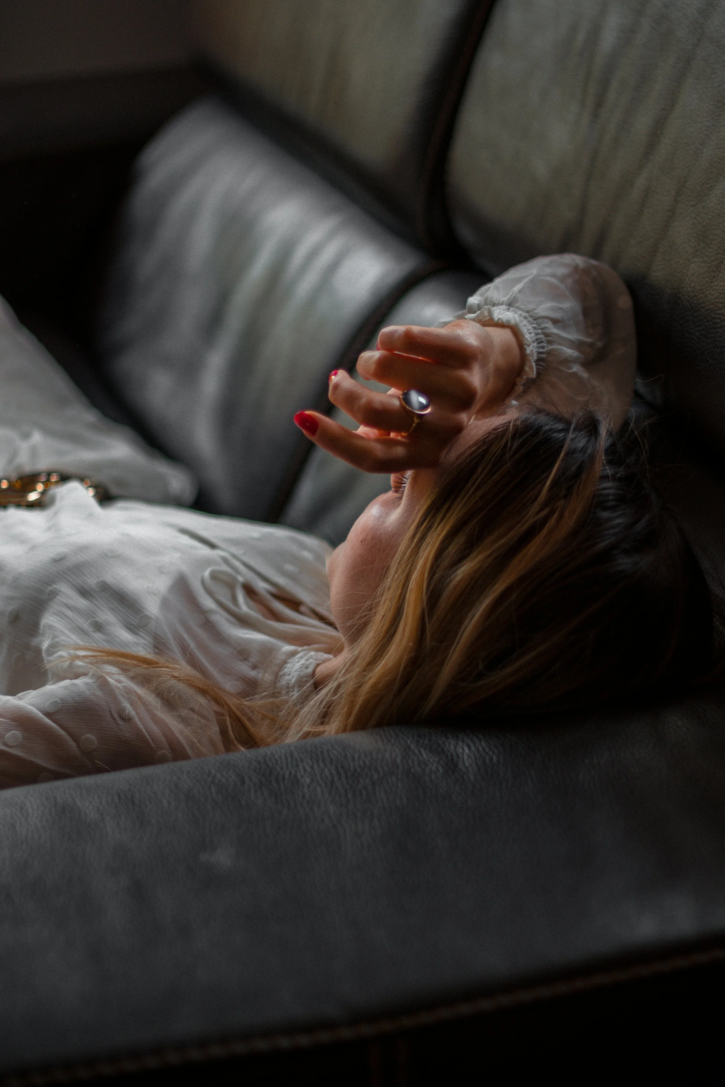 A woman with blonde hair lying on a gray sofa, resting her head on the cushion, with her eyes closed and hand resting on her forehead.