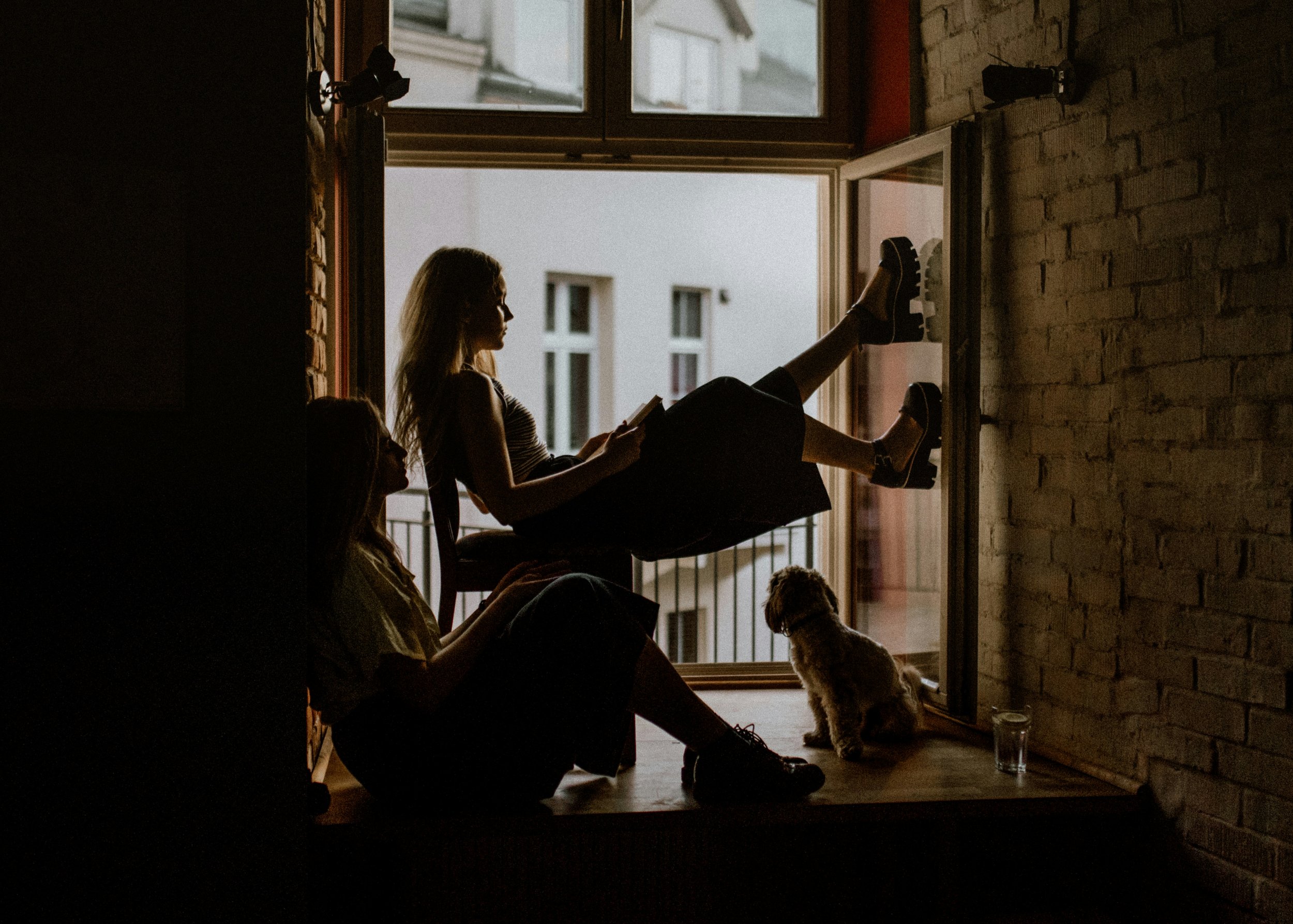 Two women and a dog sitting on a windowsill indoors, with one woman sitting and holding a book, the other woman with her legs up, looking out the window, and the dog sitting near the window, looking outside.