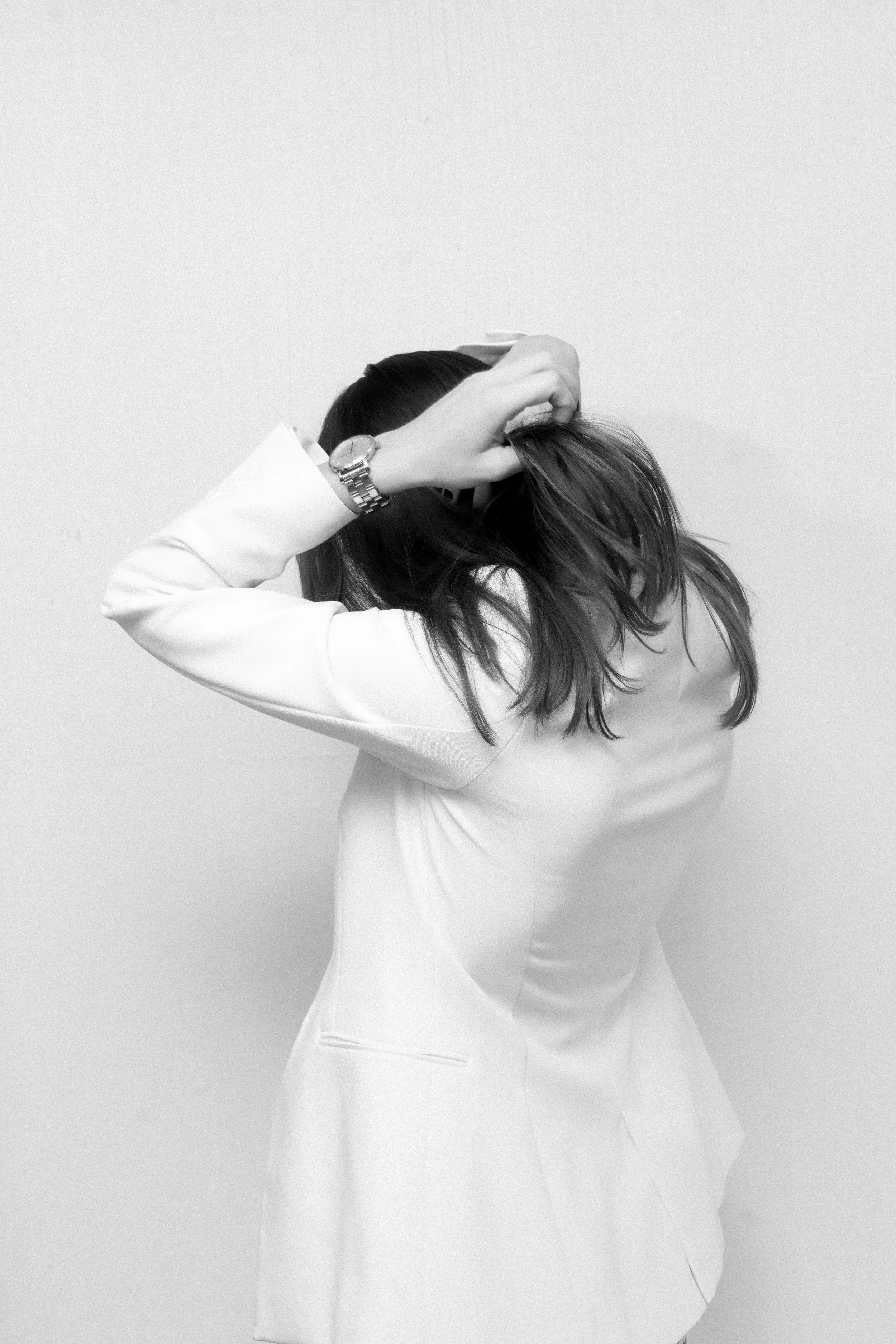 A woman adjusting her hair, wearing a white blazer and a wristwatch, standing against a plain white wall.
