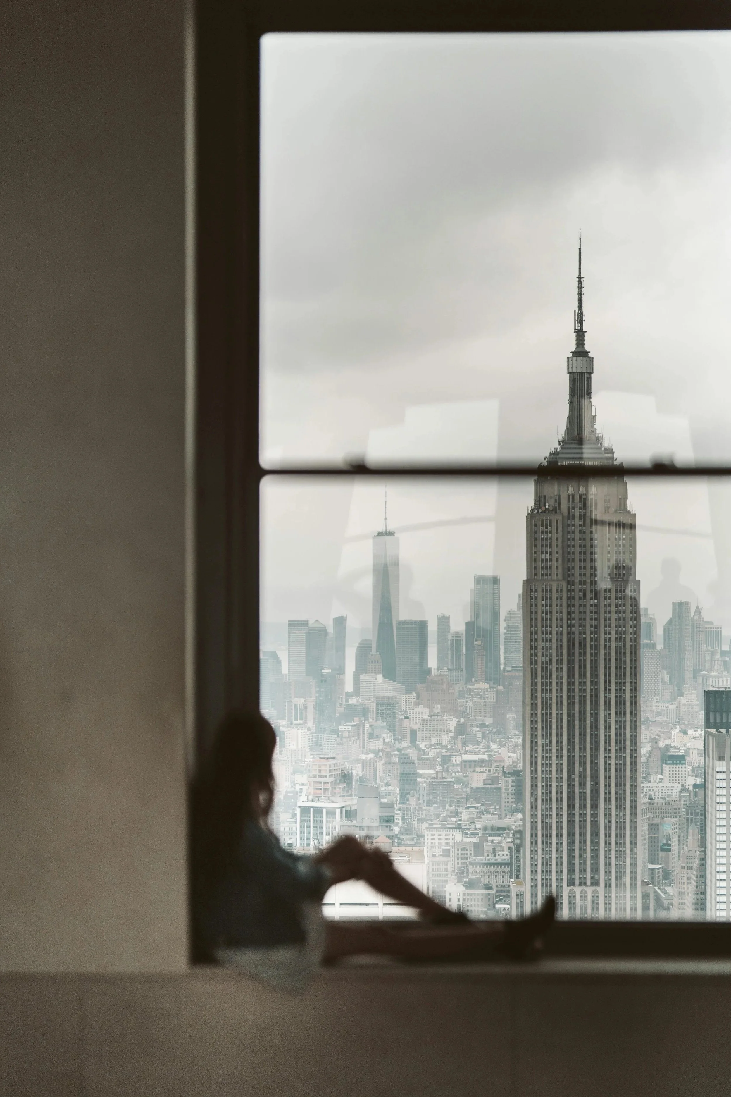 View of New York City skyline with Empire State Building and One World Trade Center through a window, with a person sitting and reading nearby.