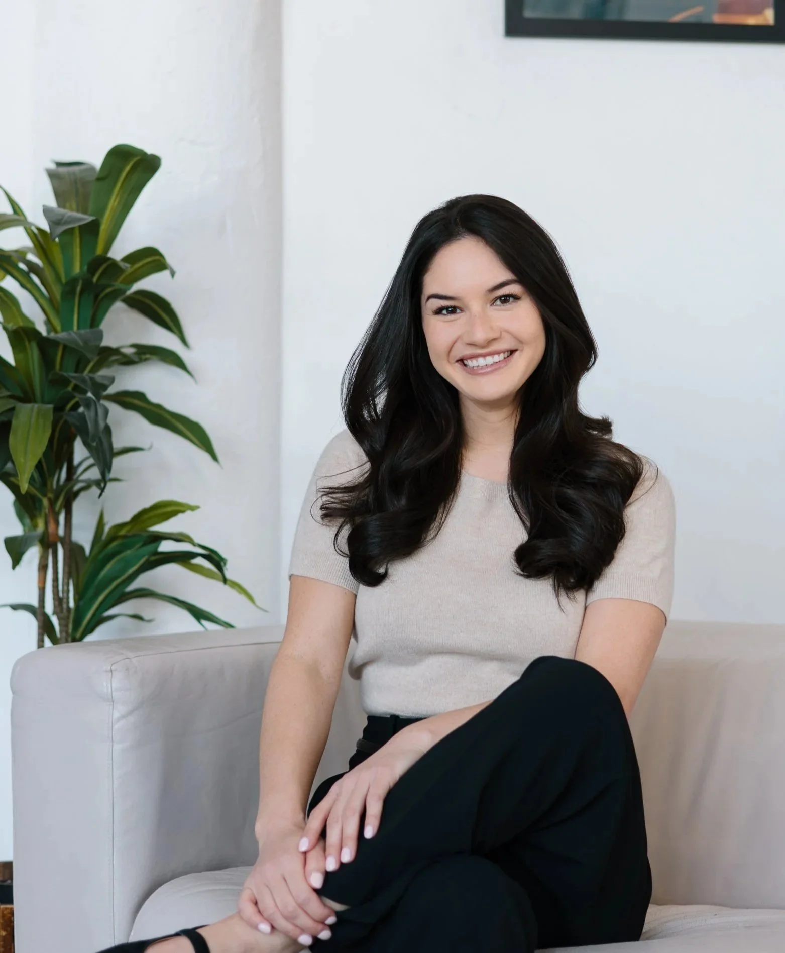 A woman with long, dark hair sitting on a light-colored sofa, smiling at the camera, with a tall green plant and a white wall in the background.